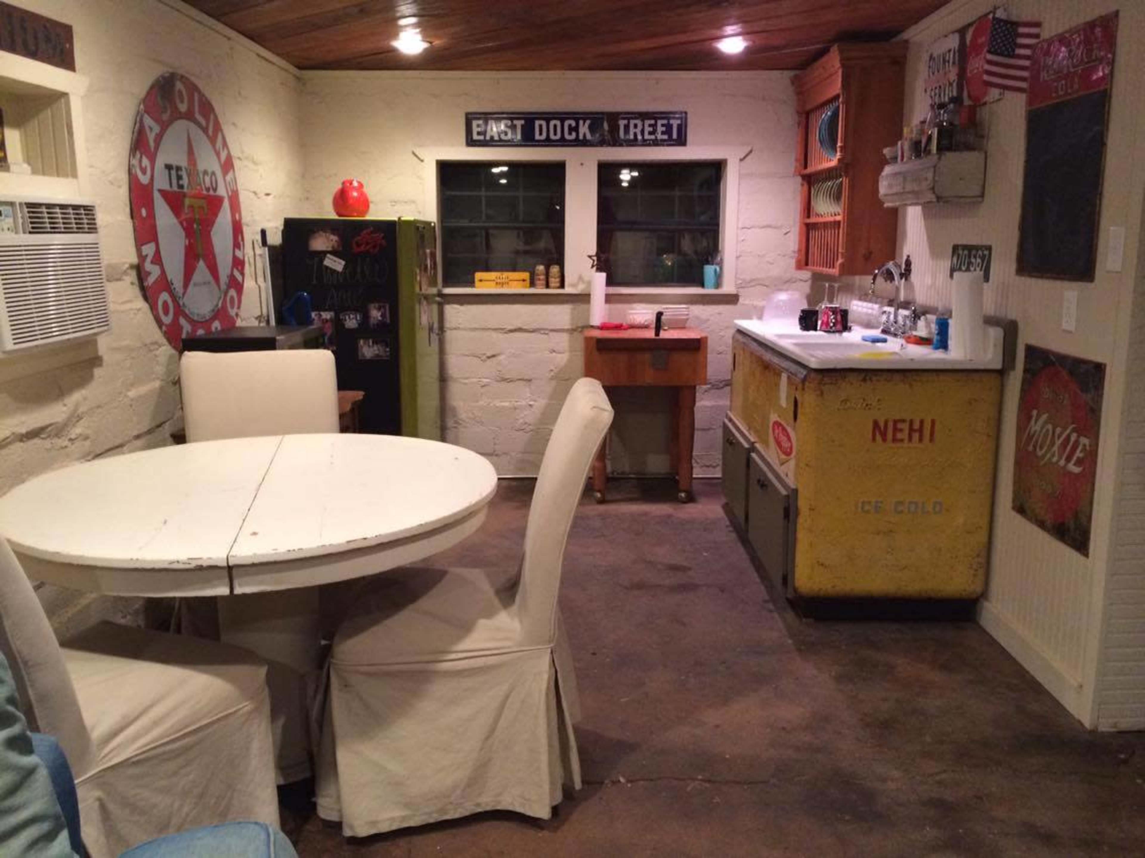 A rustic kitchen area with a round white table, four upholstered chairs, a yellow vintage ice cooler, and a window overlooking a small workspace.