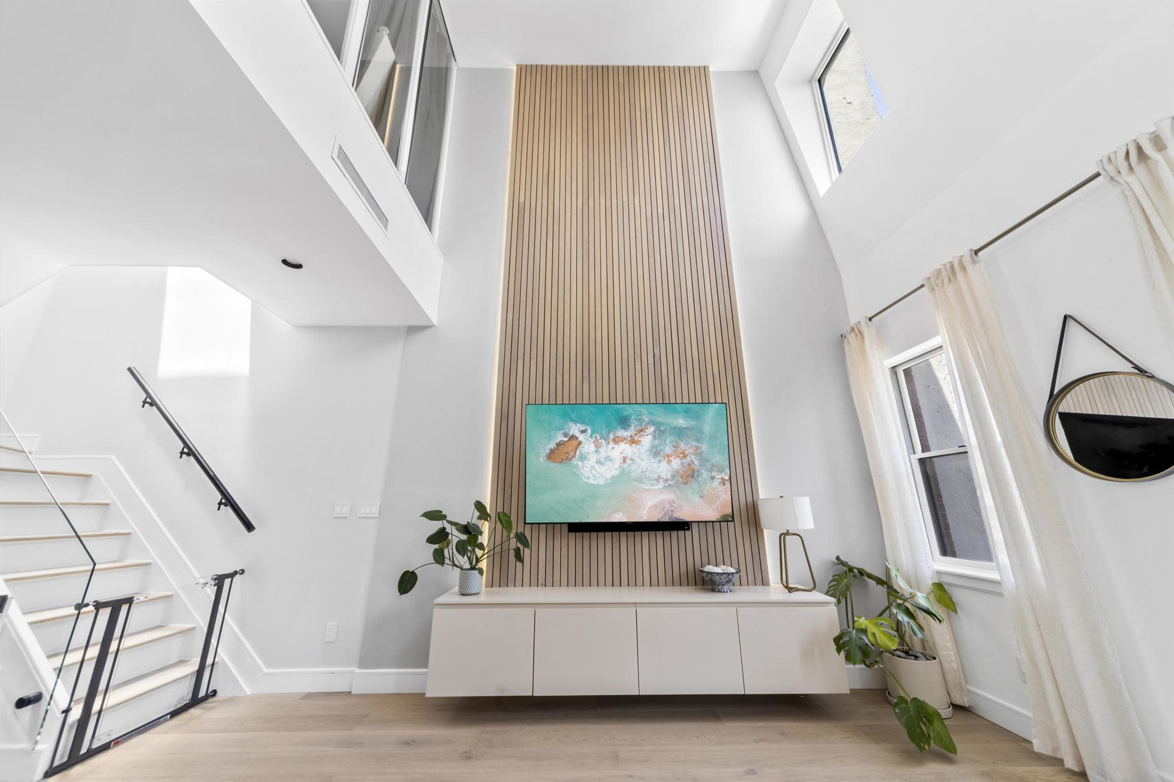 A bright, modern living room featuring a vertical wooden accent wall with a mounted television, a sleek storage cabinet below, and a staircase in the background.