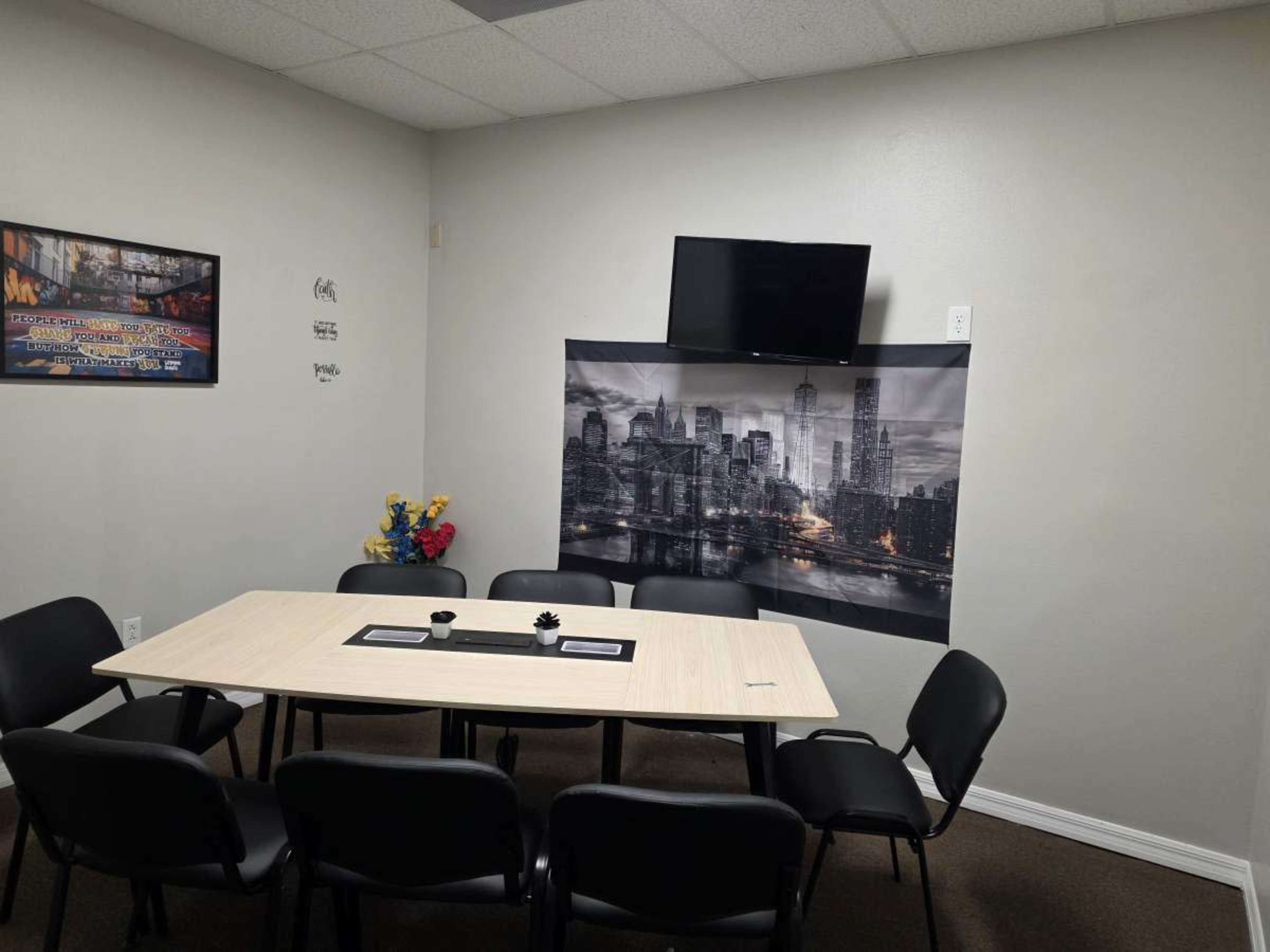 The image shows a small meeting room with a rectangular table surrounded by black chairs, a wall-mounted television, and a large black-and-white cityscape mural.