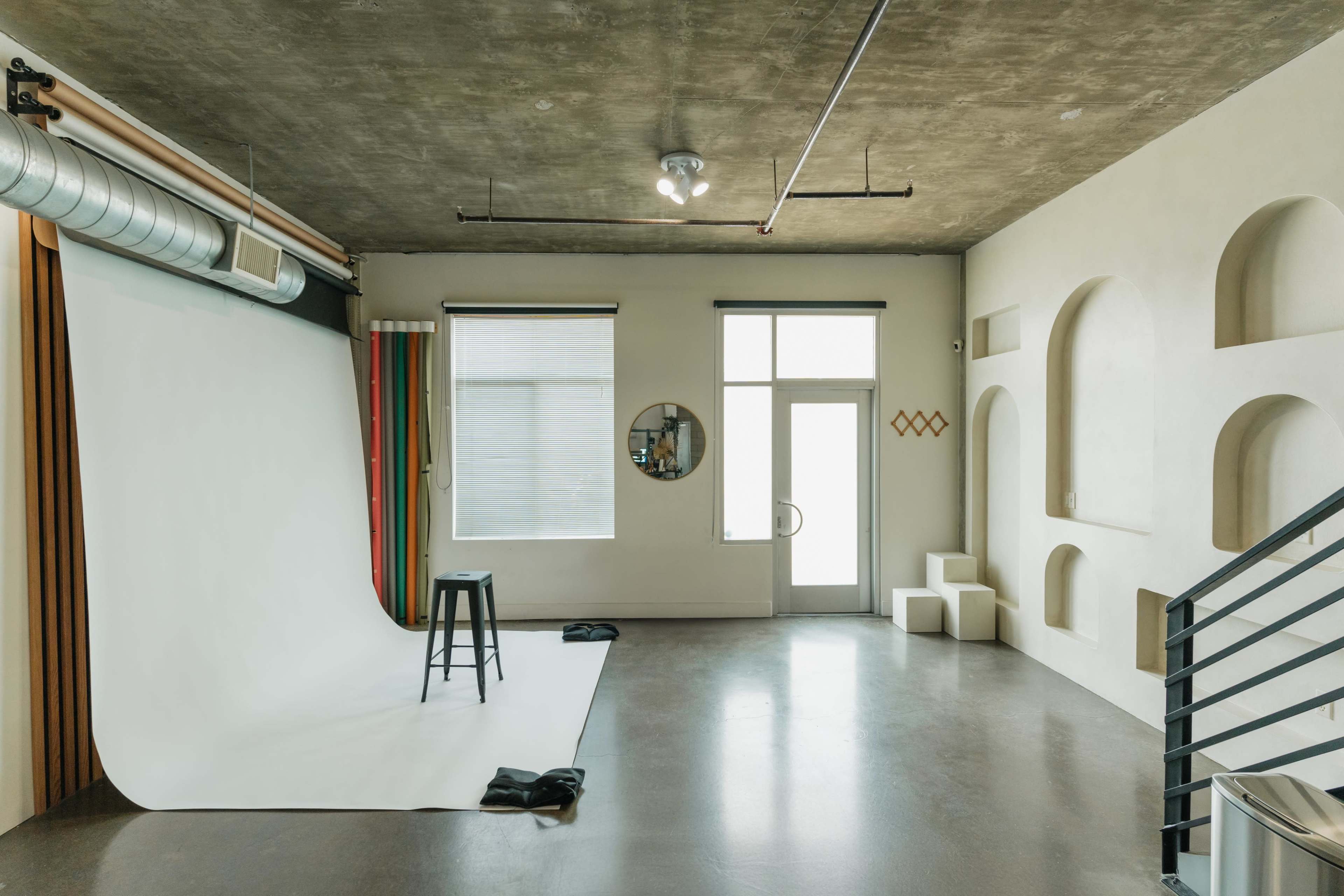 The image shows a modern studio space with a white backdrop, a black stool, and architectural details including arched niches and large windows.