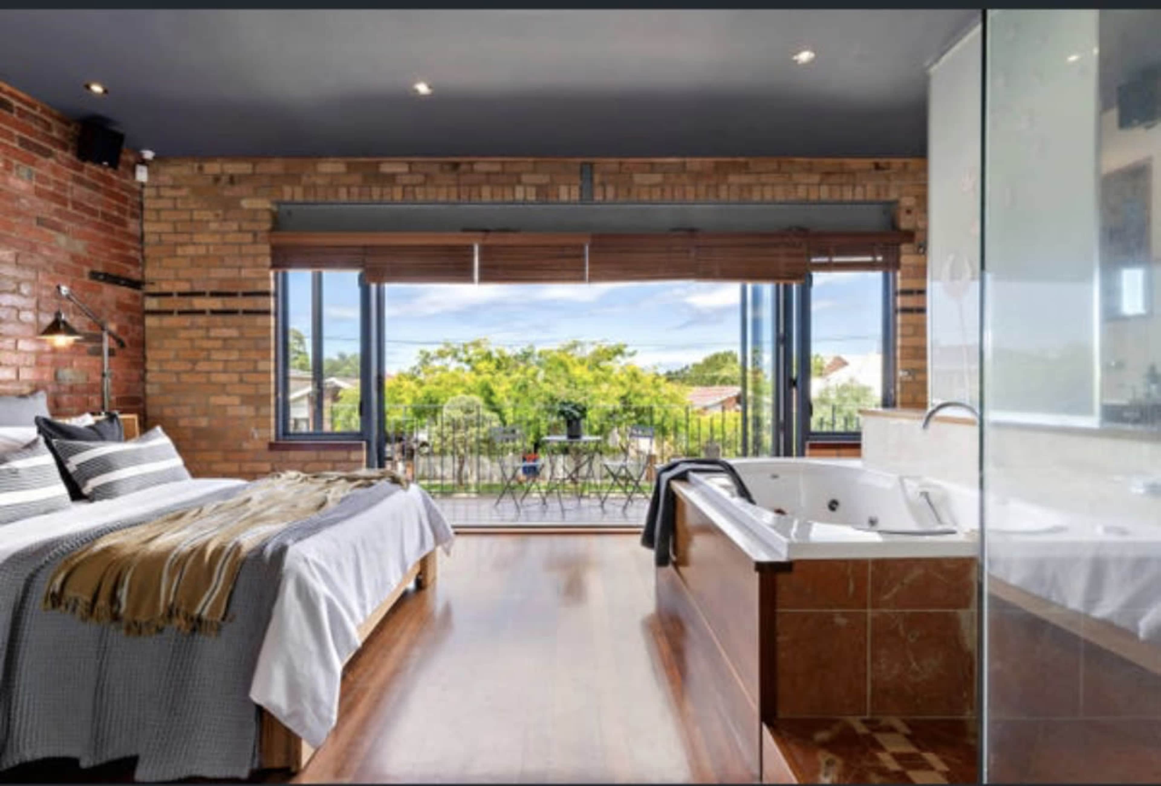A modern bedroom with a large bed, a jacuzzi tub, and wide windows offering a view of an outdoor balcony.