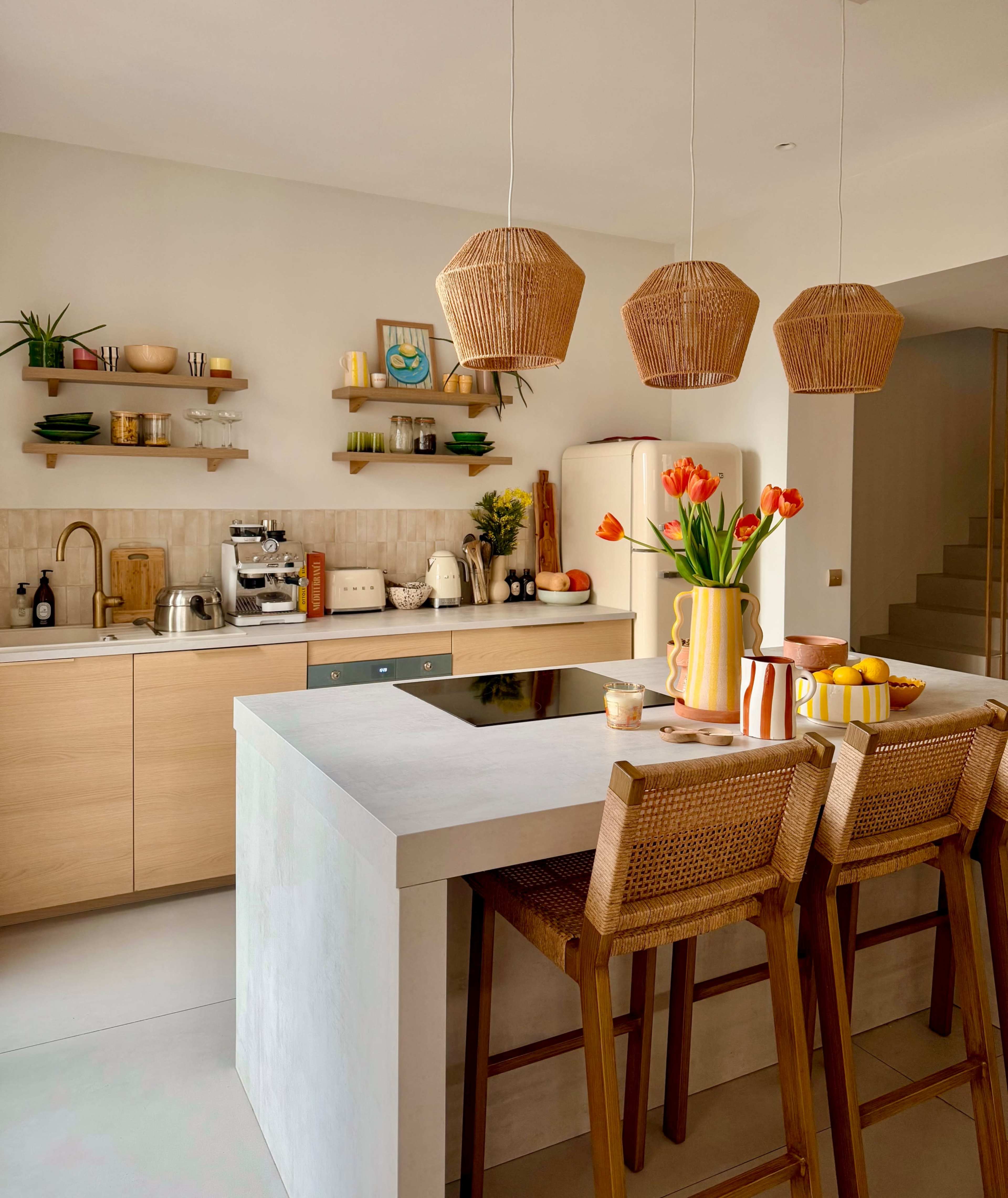 The image shows a modern kitchen with wooden cabinetry, a large island with bar stools, and decorative items including flowers and fruit.
