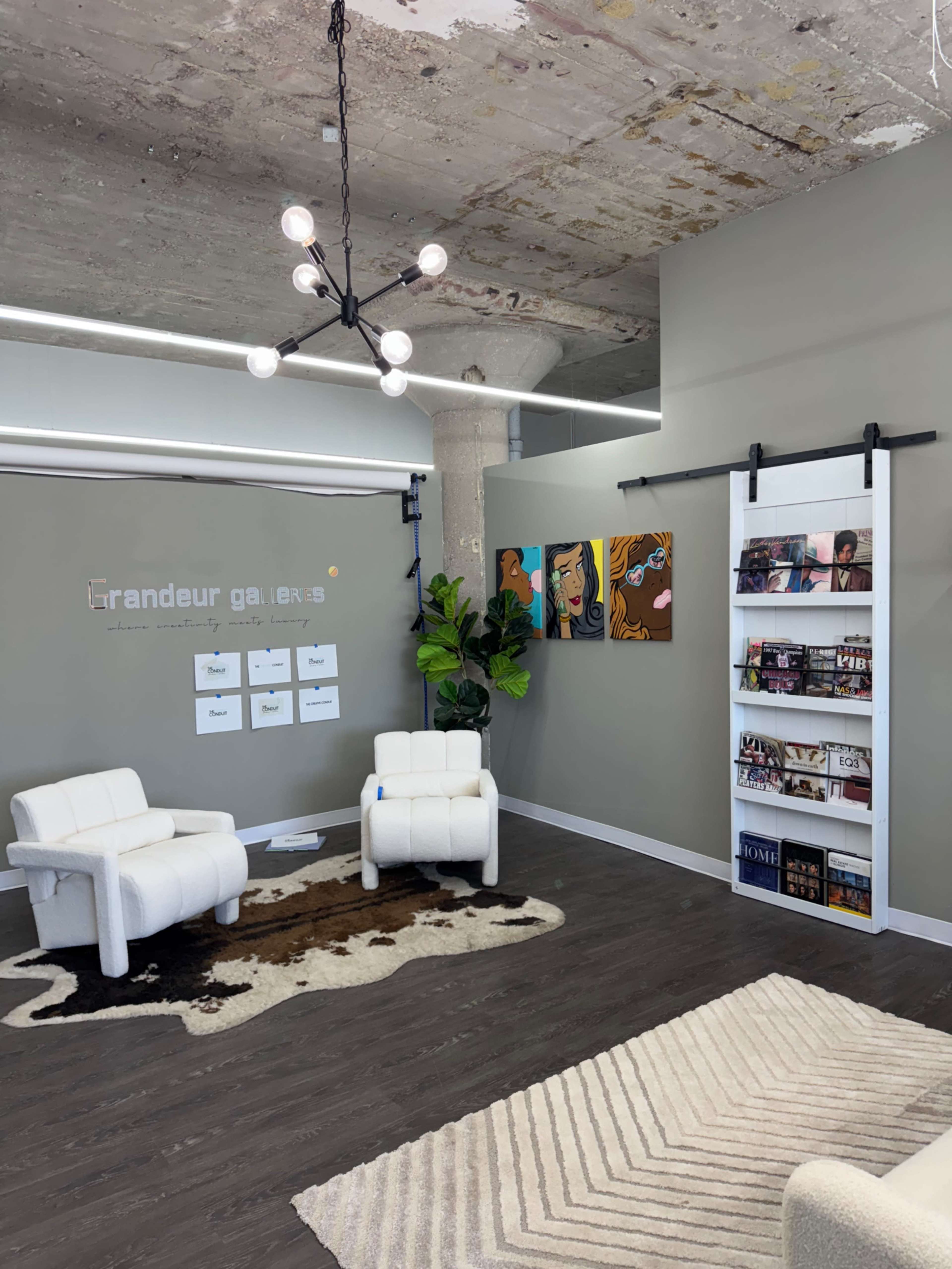The image shows a modern interior space with two white chairs, a patterned rug, a wall-mounted magazine rack, and artistic wall decor including framed artworks and a sign that reads "grandeur gallery."