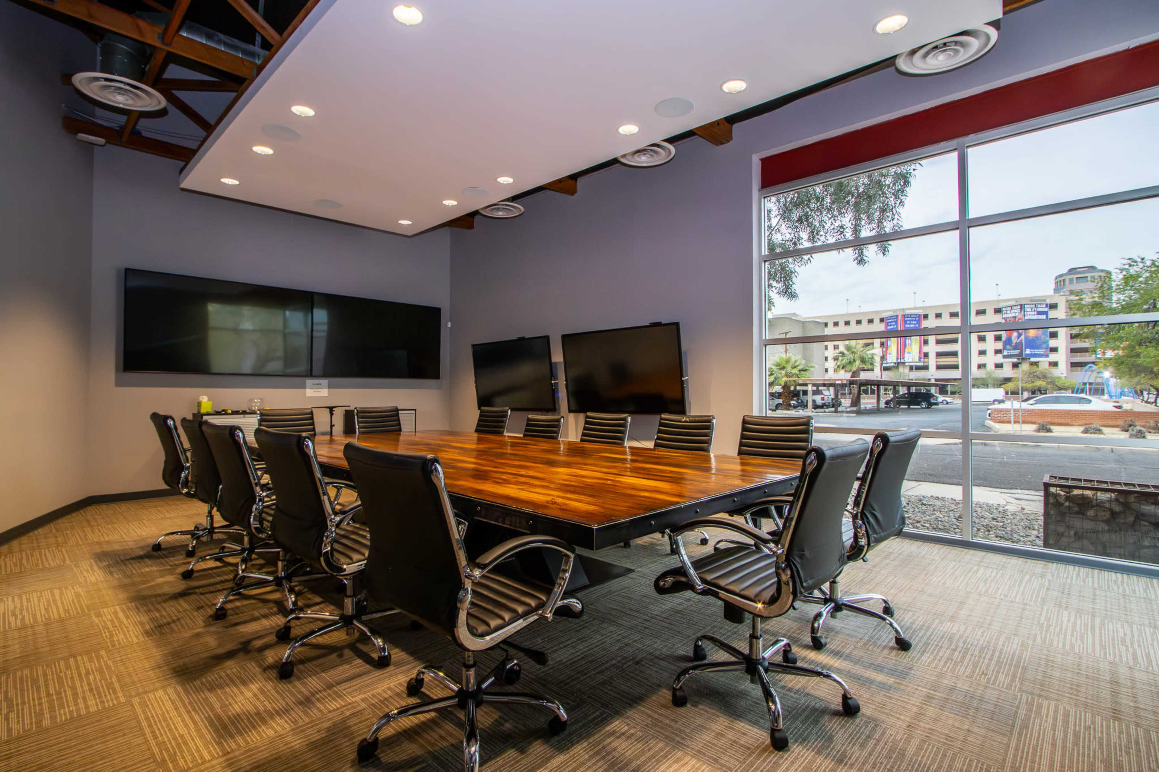 A conference room features a long wooden table surrounded by black leather chairs and multiple large screens on the walls.