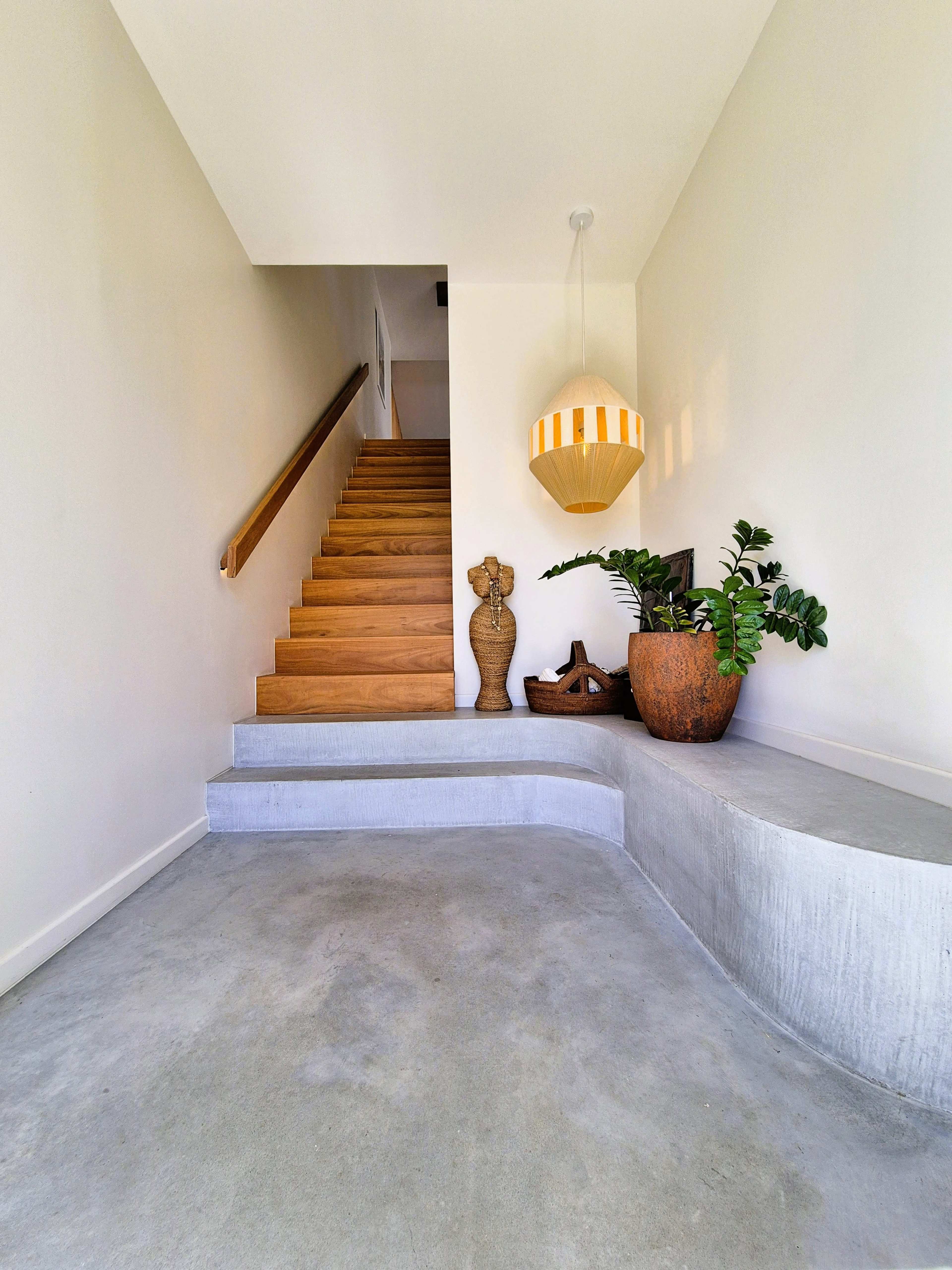 The image shows a modern foyer with a staircase leading to an upper level, featuring a large potted plant and decorative sculptures.