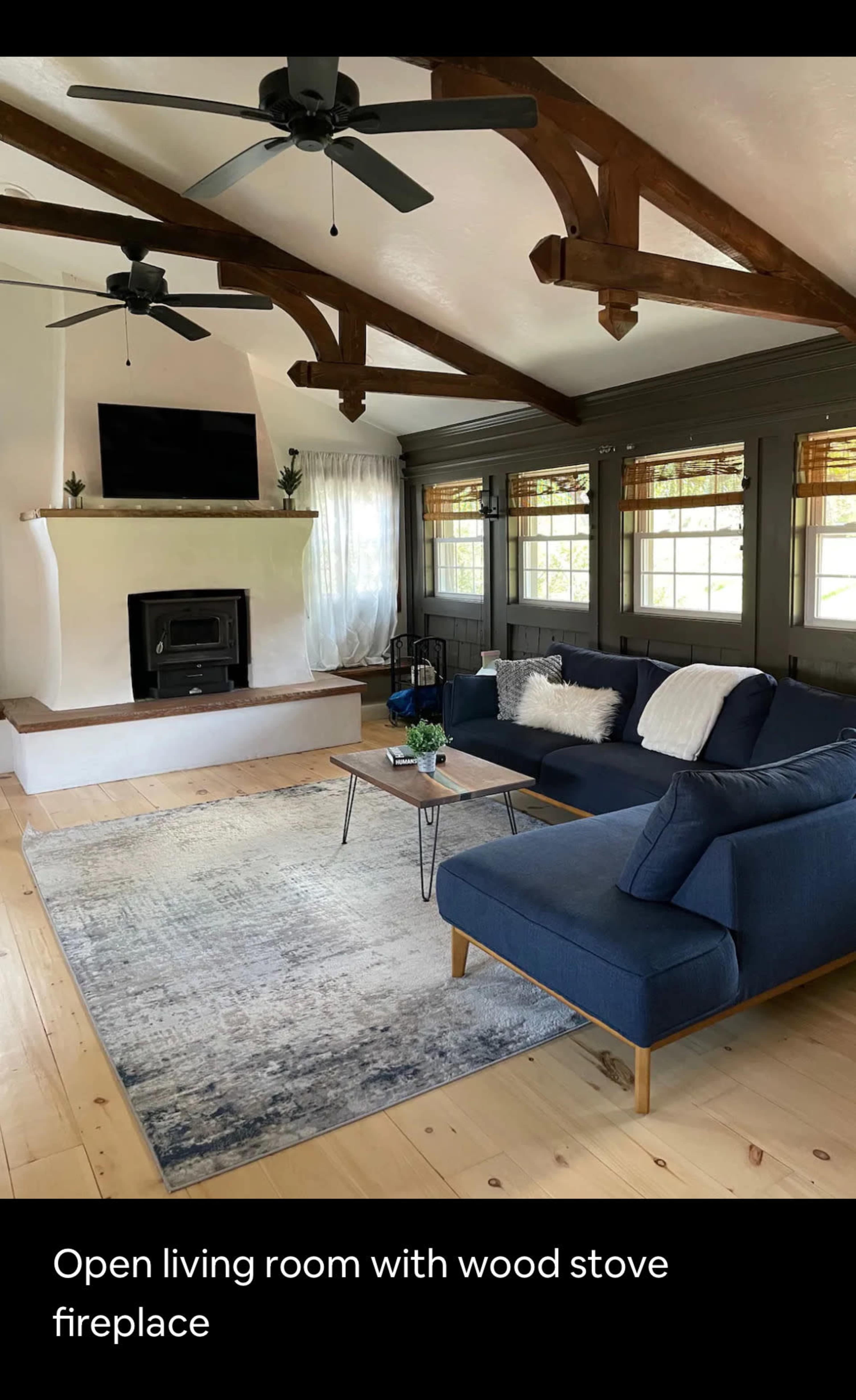 A spacious living room features a wood stove fireplace, large windows, and exposed wooden beams with a navy blue sectional sofa and a coffee table.