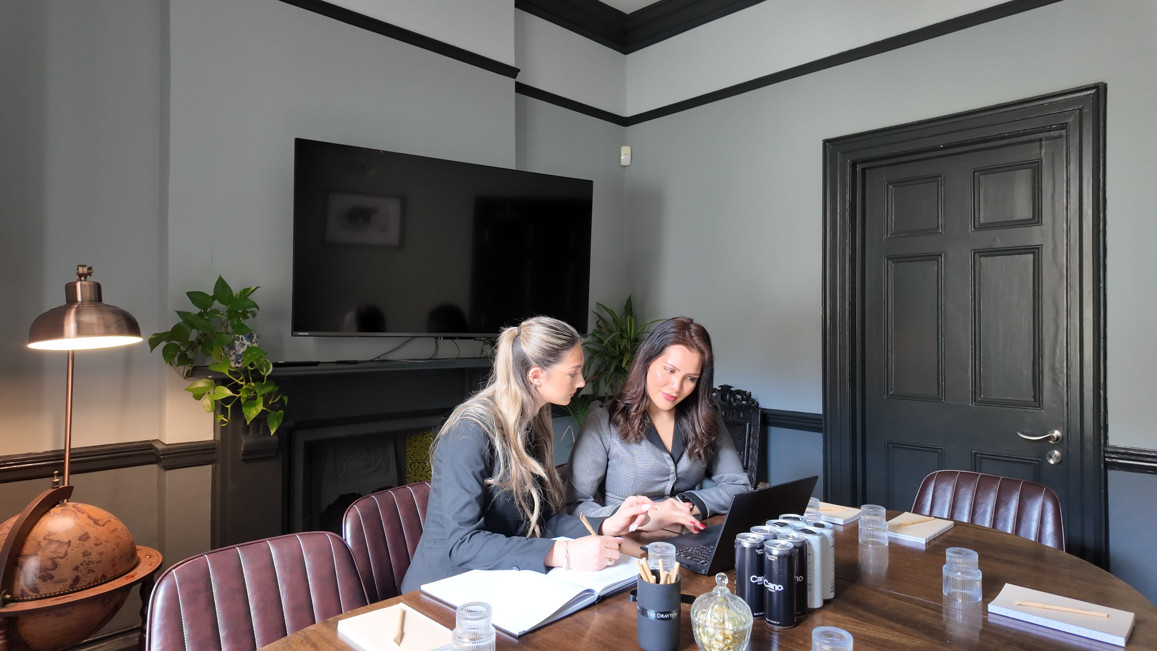 Two women are sitting at a conference table, collaborating on a laptop in a professional meeting space with a large television screen and decorative plants in the background.