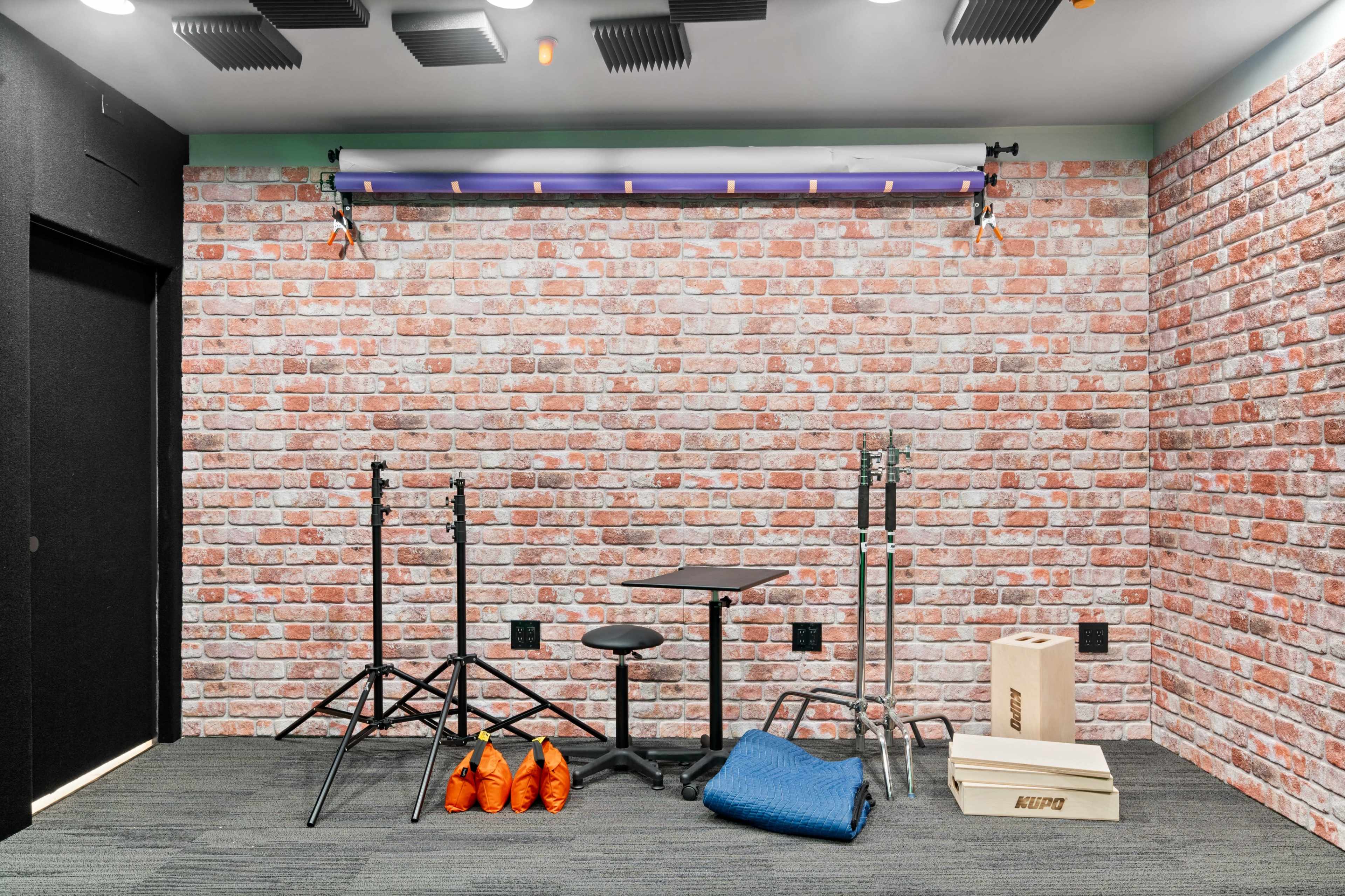 A studio space with a brick wall, a stool, lighting equipment stands, a blue blanket, and wooden boxes on the floor.
