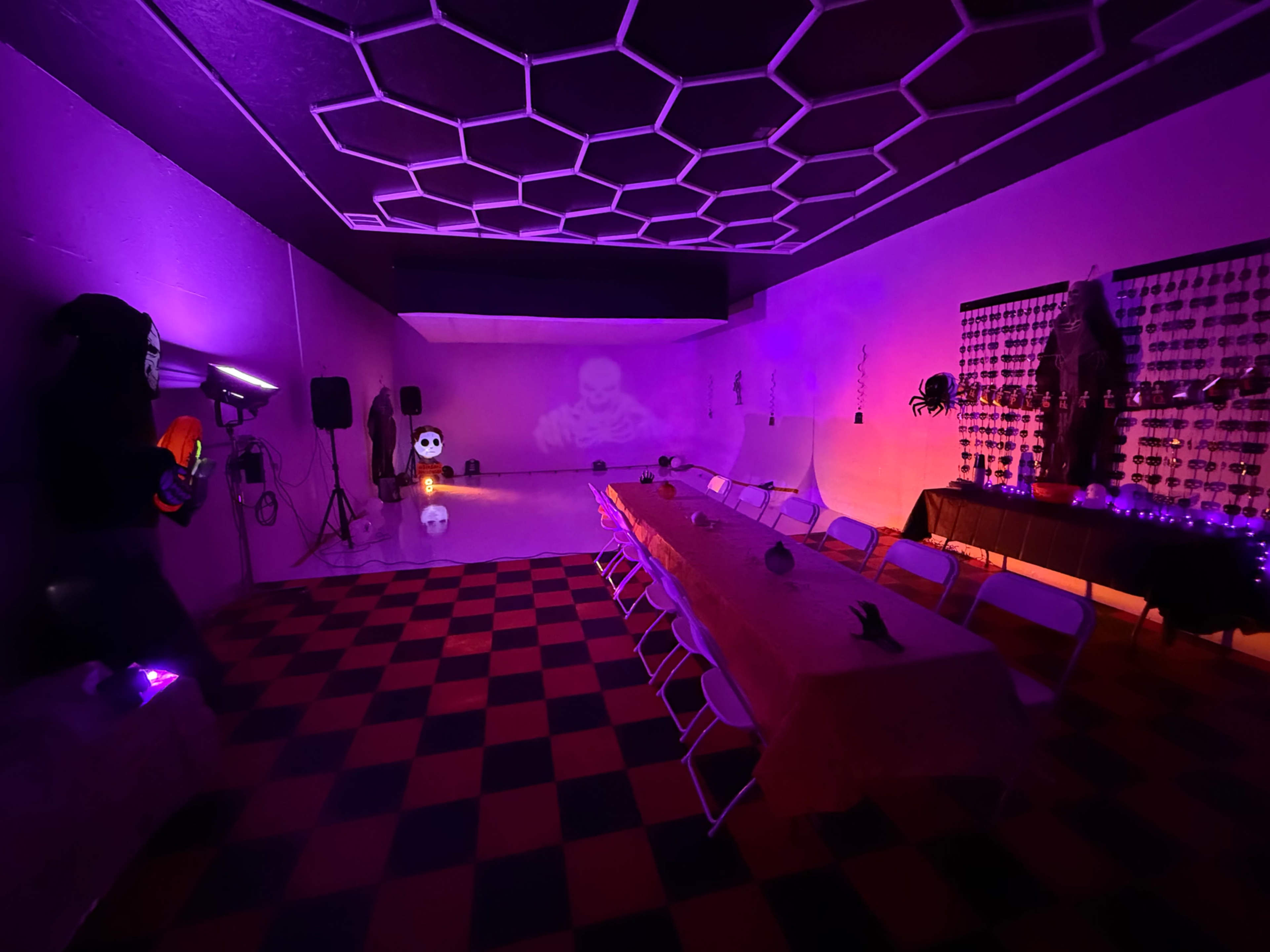 A dimly lit room with purple lighting features a long table set for a gathering, surrounded by Halloween decorations including skeletons and spiders.