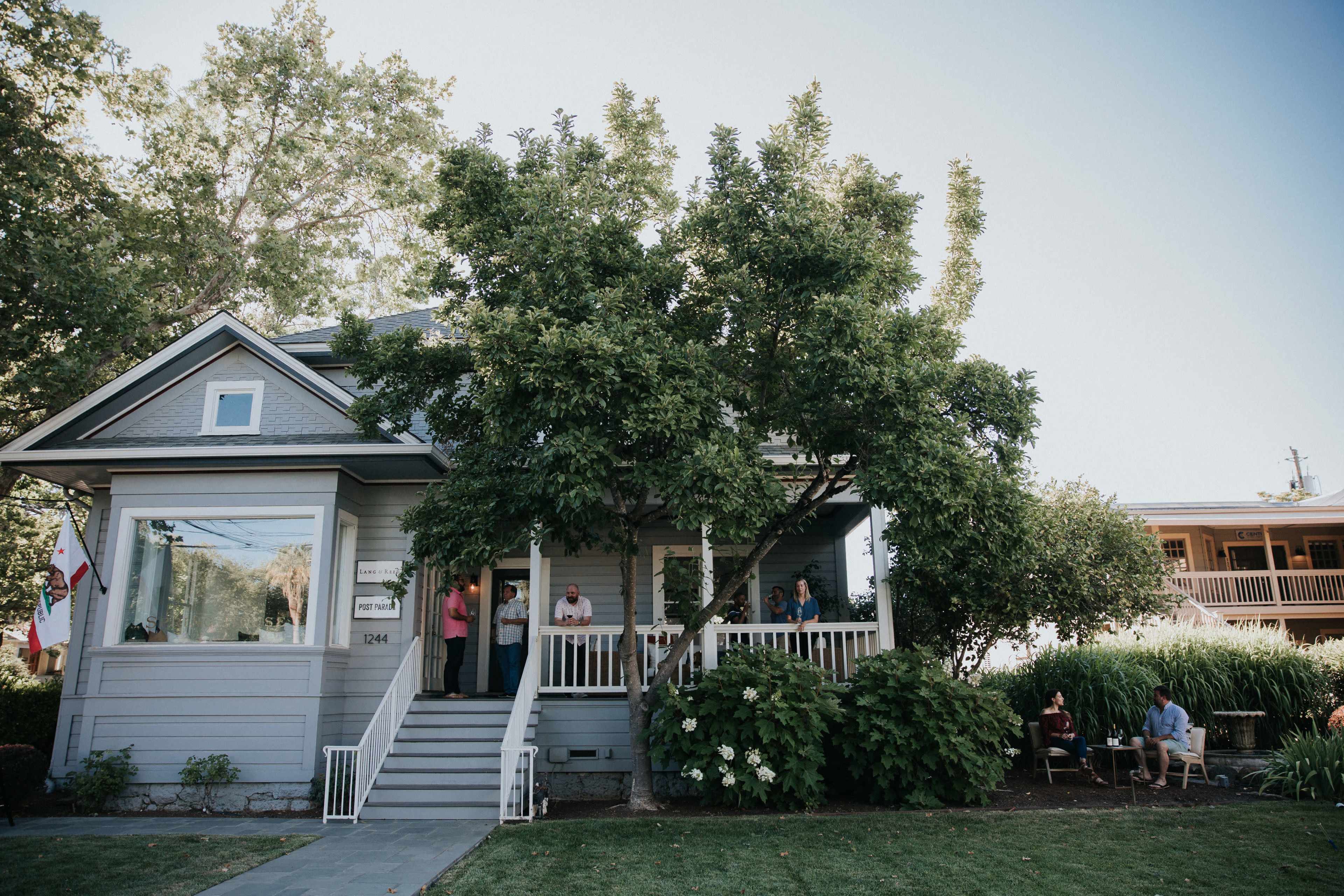 A house with a front porch and several people gathered outside, surrounded by trees and greenery.