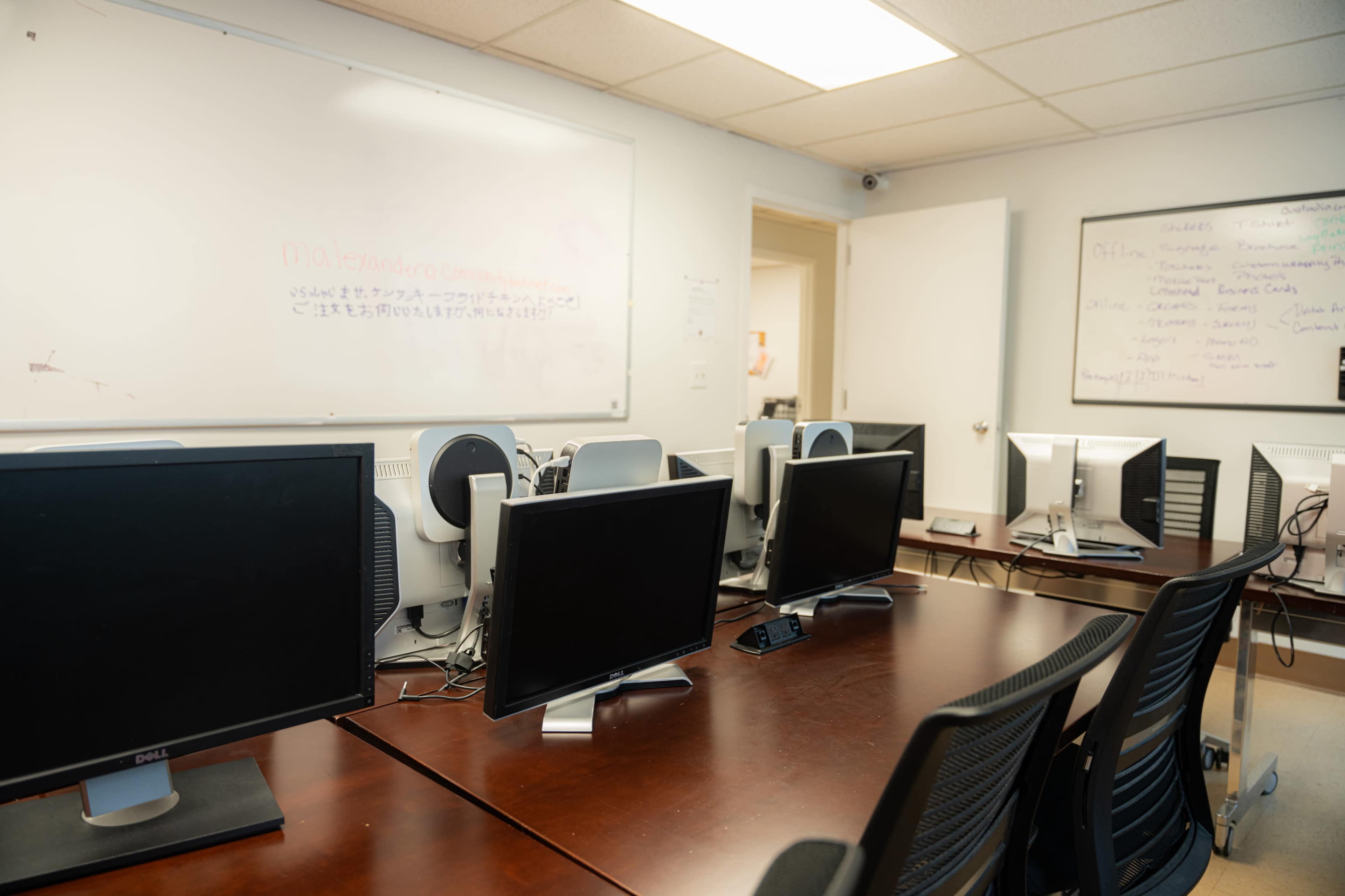 A classroom or office space with several desktop computers arranged on wooden tables, a whiteboard in the background, and a door leading to another area.
