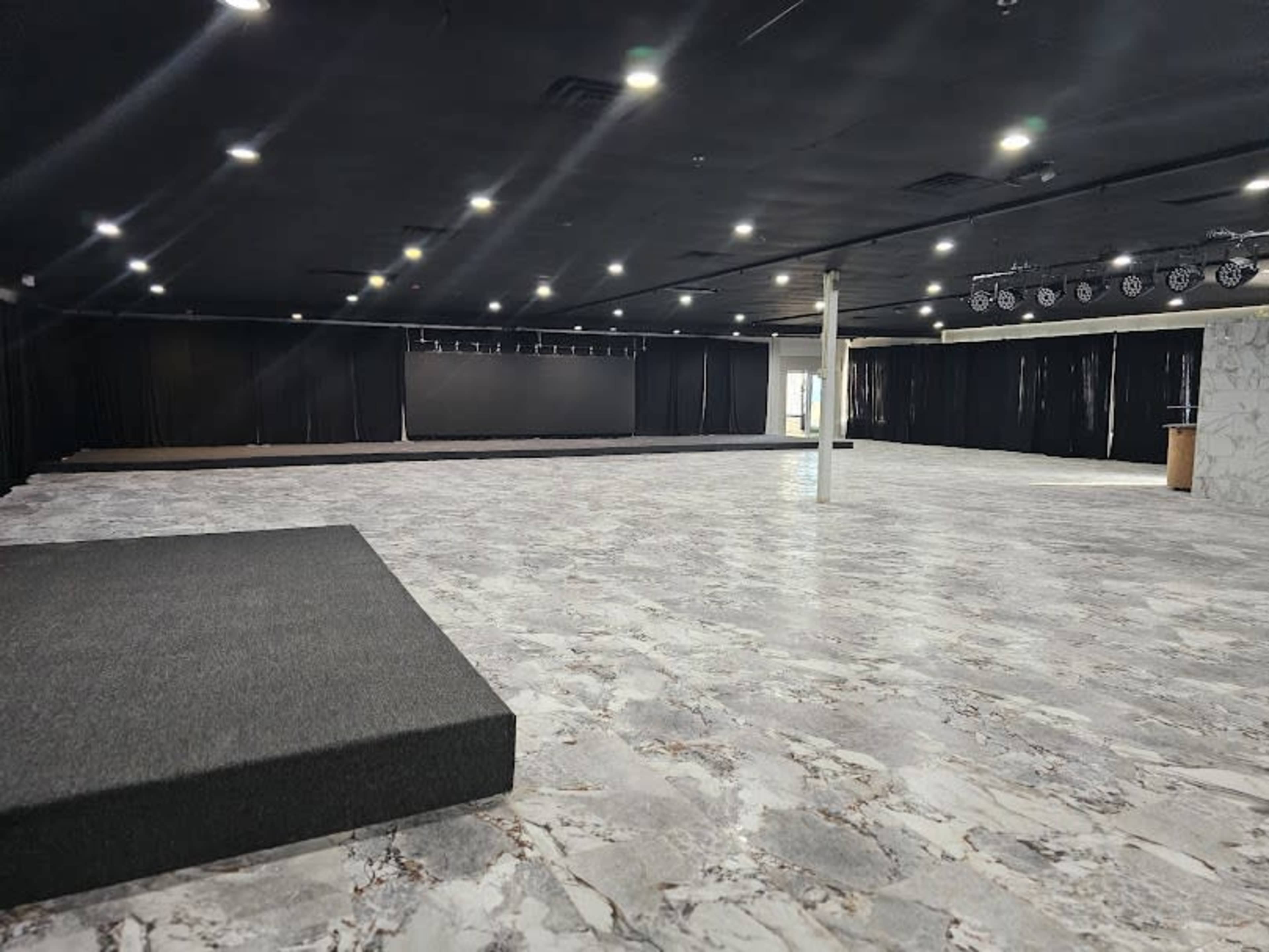 The image shows a spacious, empty room with a patterned floor, black drapes along the walls, and a small raised platform on one side.