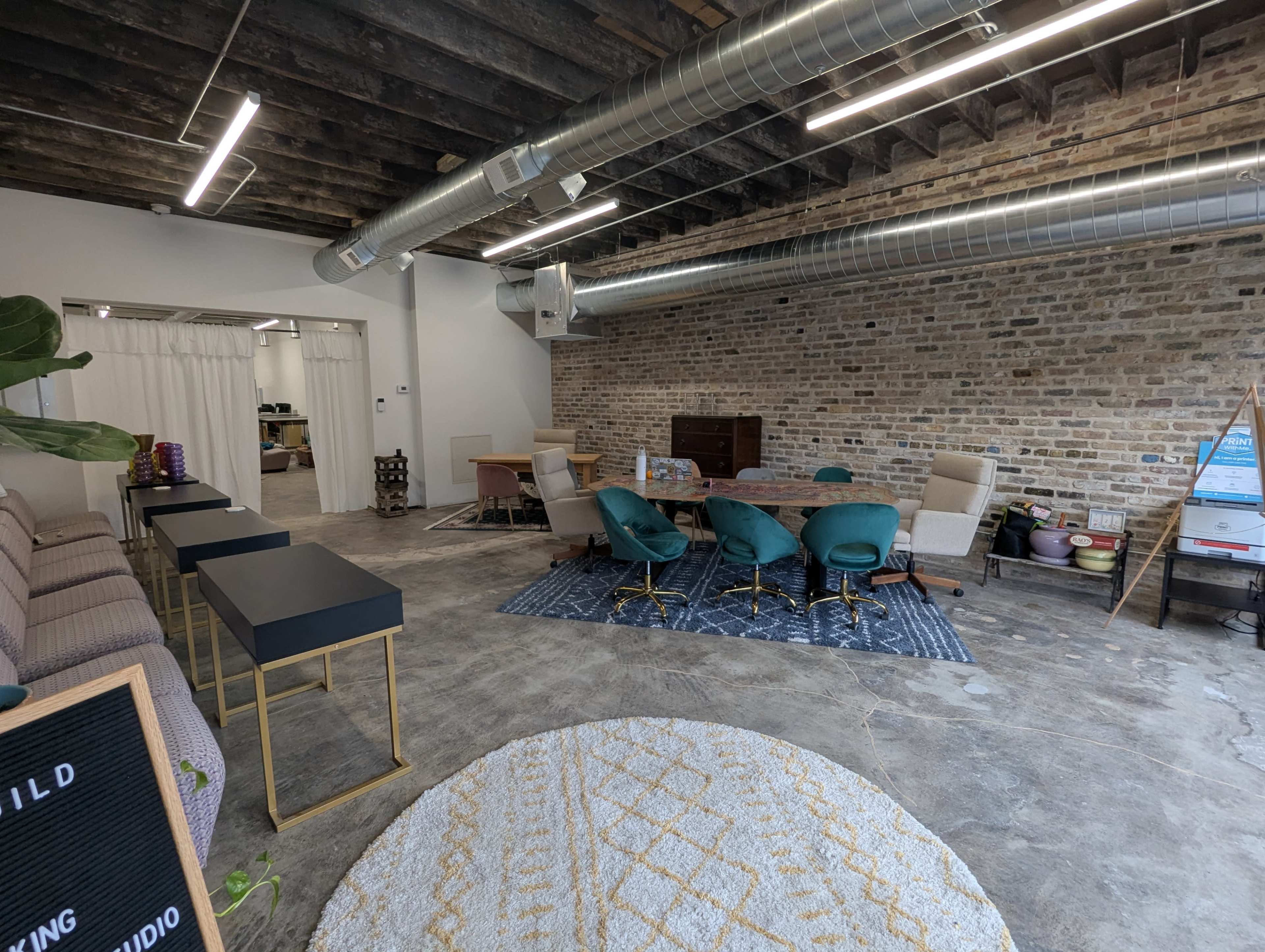The image shows a modern interior space with a mix of seating options, a dining table, exposed brick walls, and industrial-style ductwork.