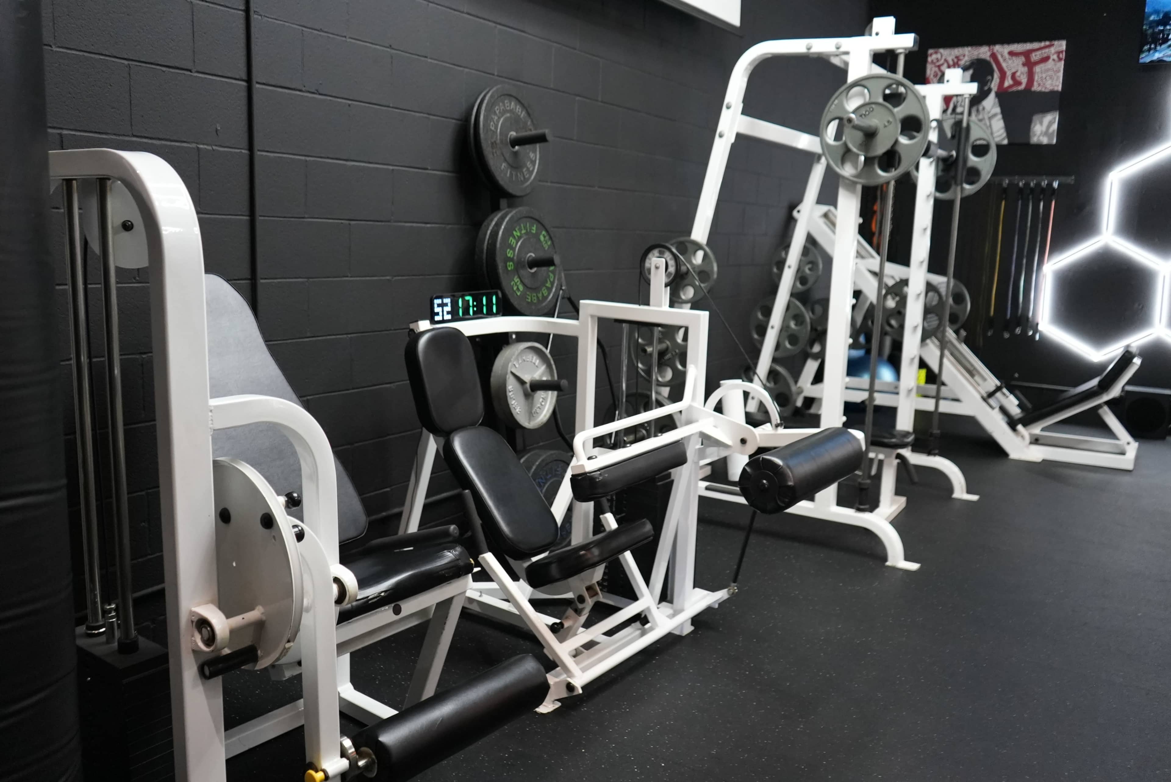 A gym interior with various weightlifting machines, including a leg press and a cable machine, positioned against a black wall.