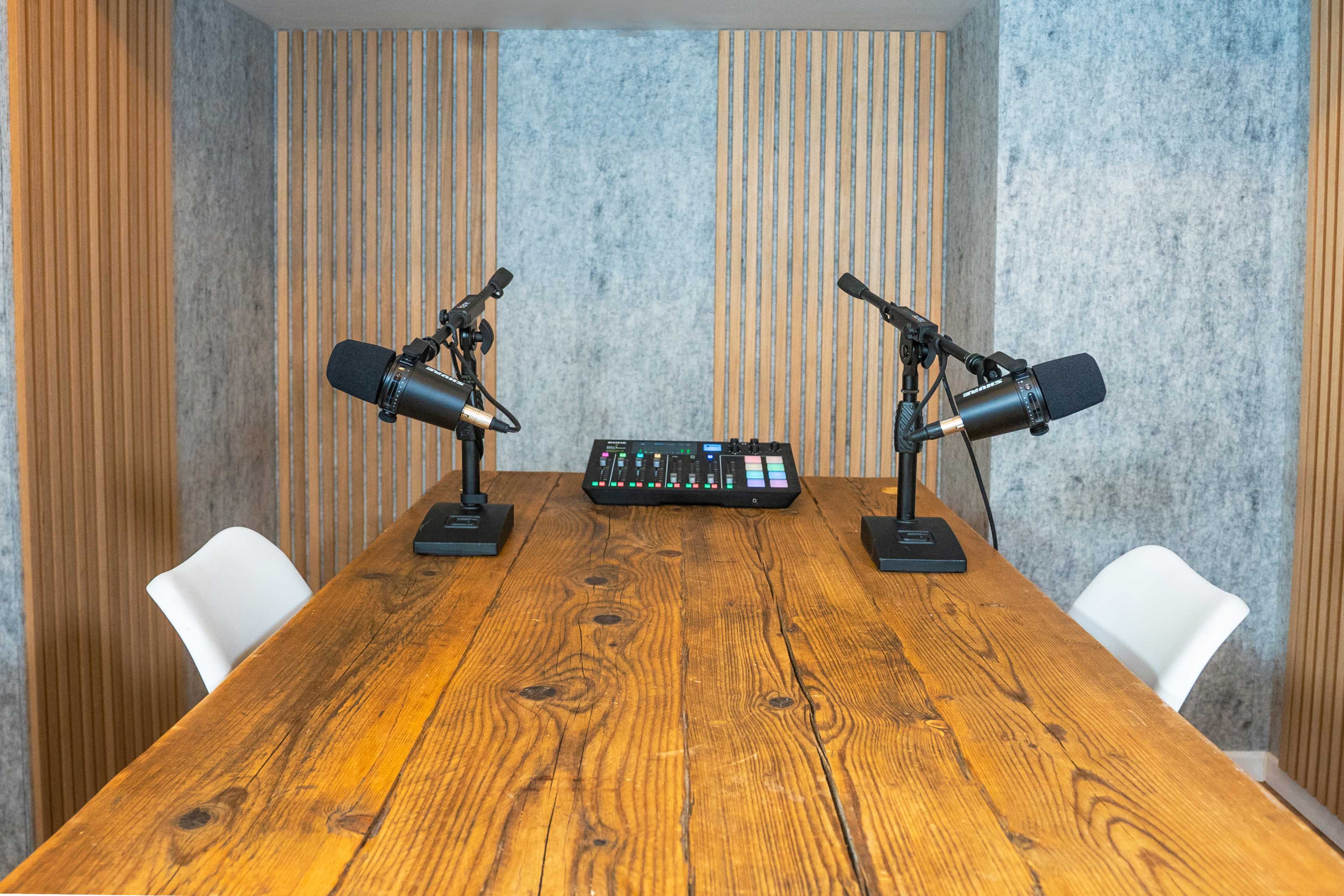 A wooden table with two microphones on stands is positioned in front of a soundboard in a room with textured walls and vertical wood paneling.