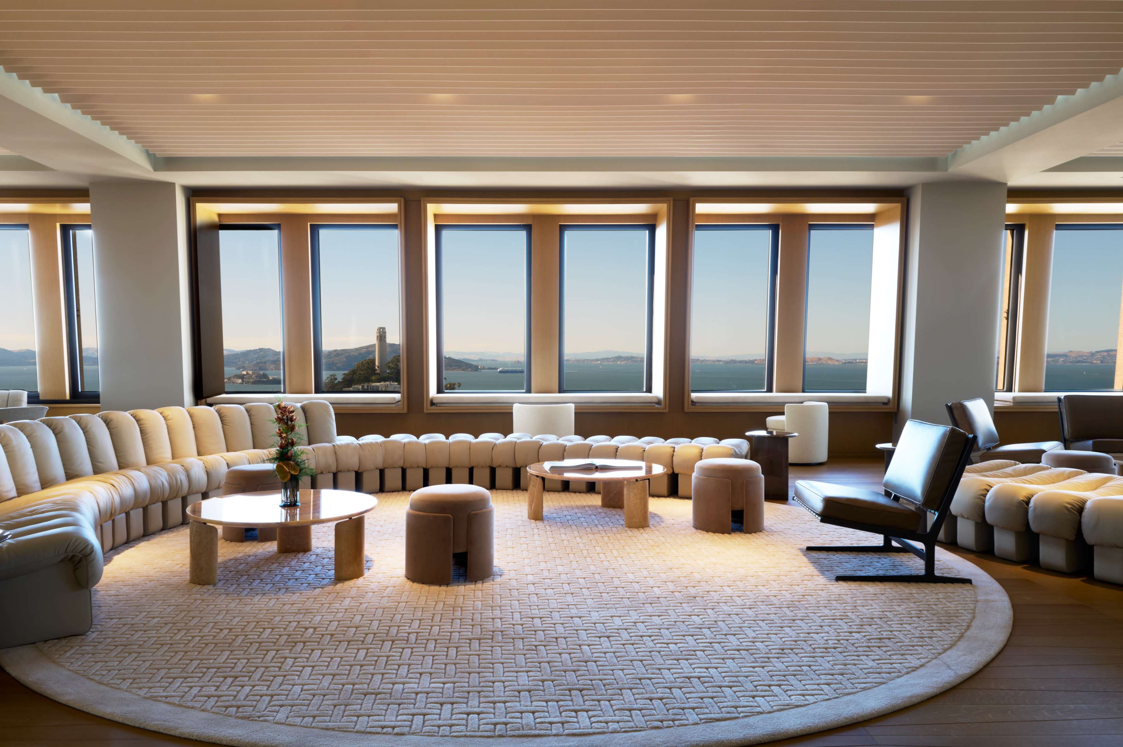 The image shows a modern living room with a curved beige sofa, several round tables, and large windows offering a view of the water and distant hills.