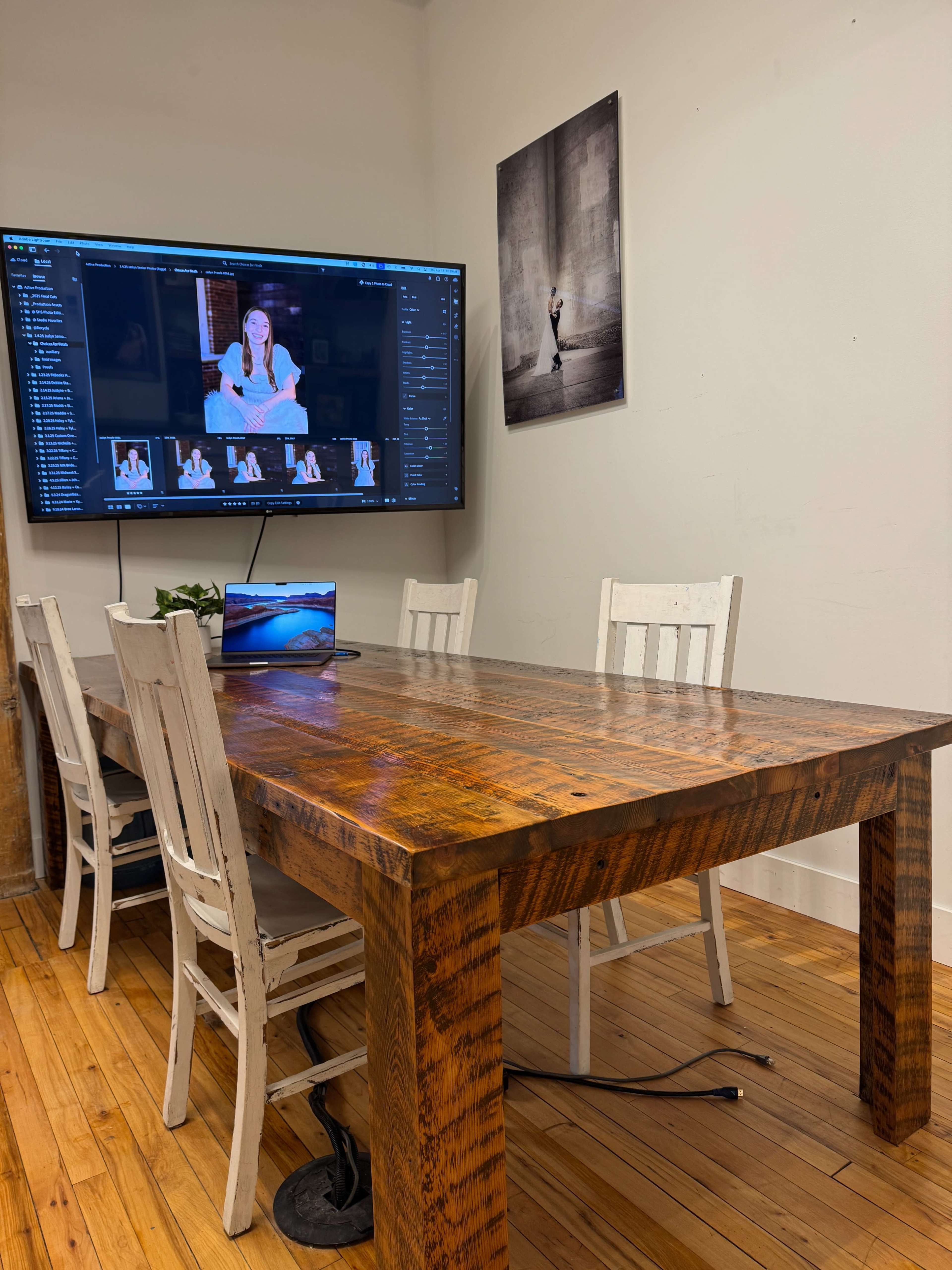 A wooden table is surrounded by white chairs in a room with a large screen displaying a photo editing interface and a framed image on the wall.