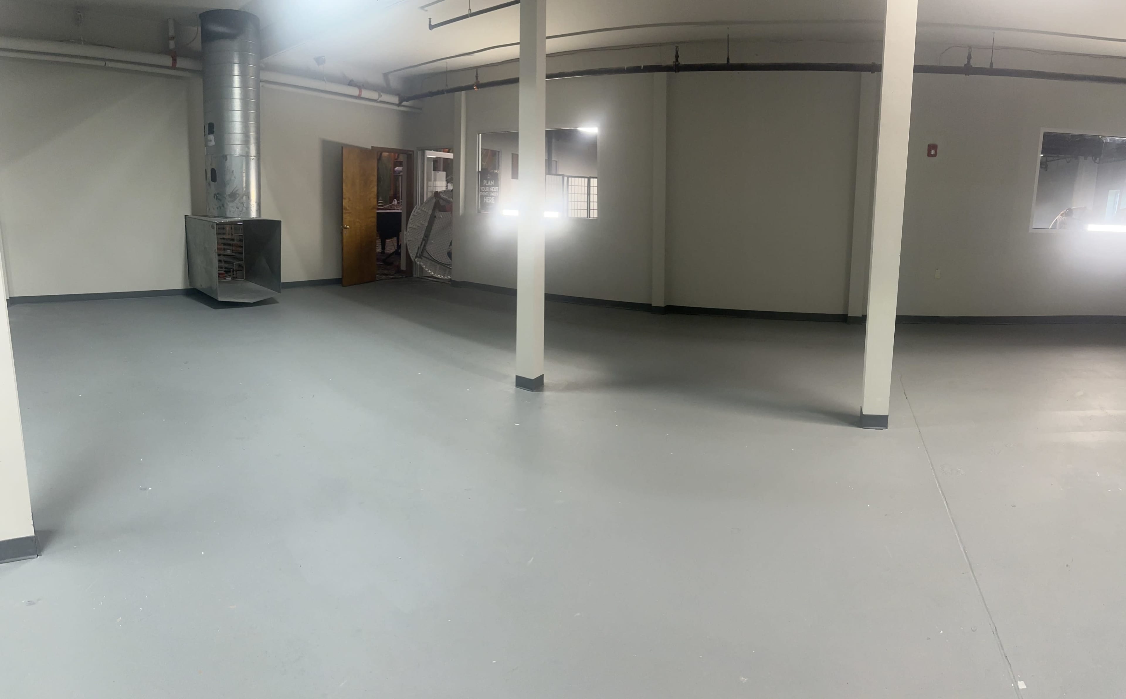 The image shows a spacious, empty room with gray flooring, several white pillars, and a doorway leading to another area.