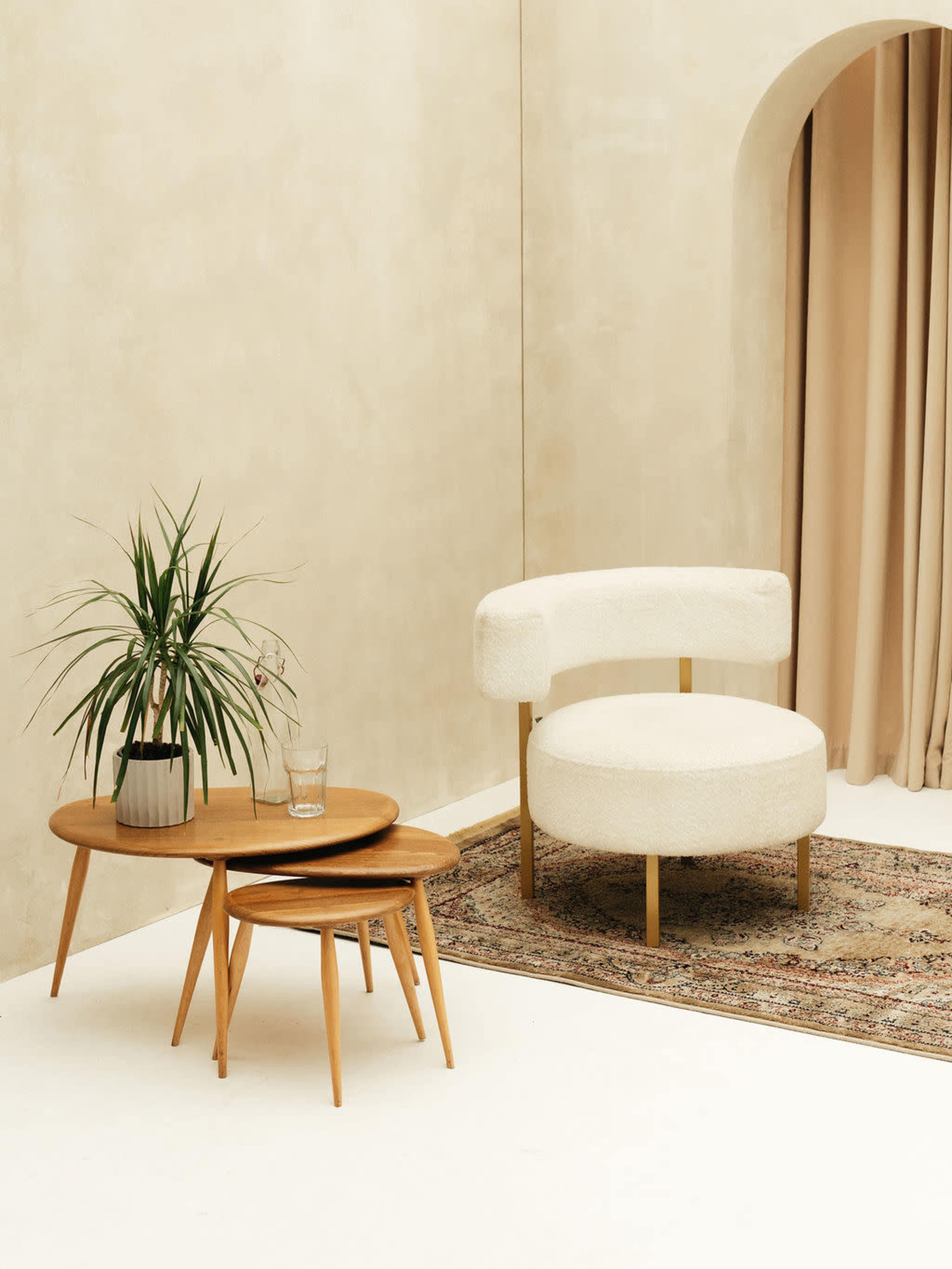 A round, cream-colored chair sits beside two wooden coffee tables on a patterned rug in a minimalist room with beige walls and draped curtains.