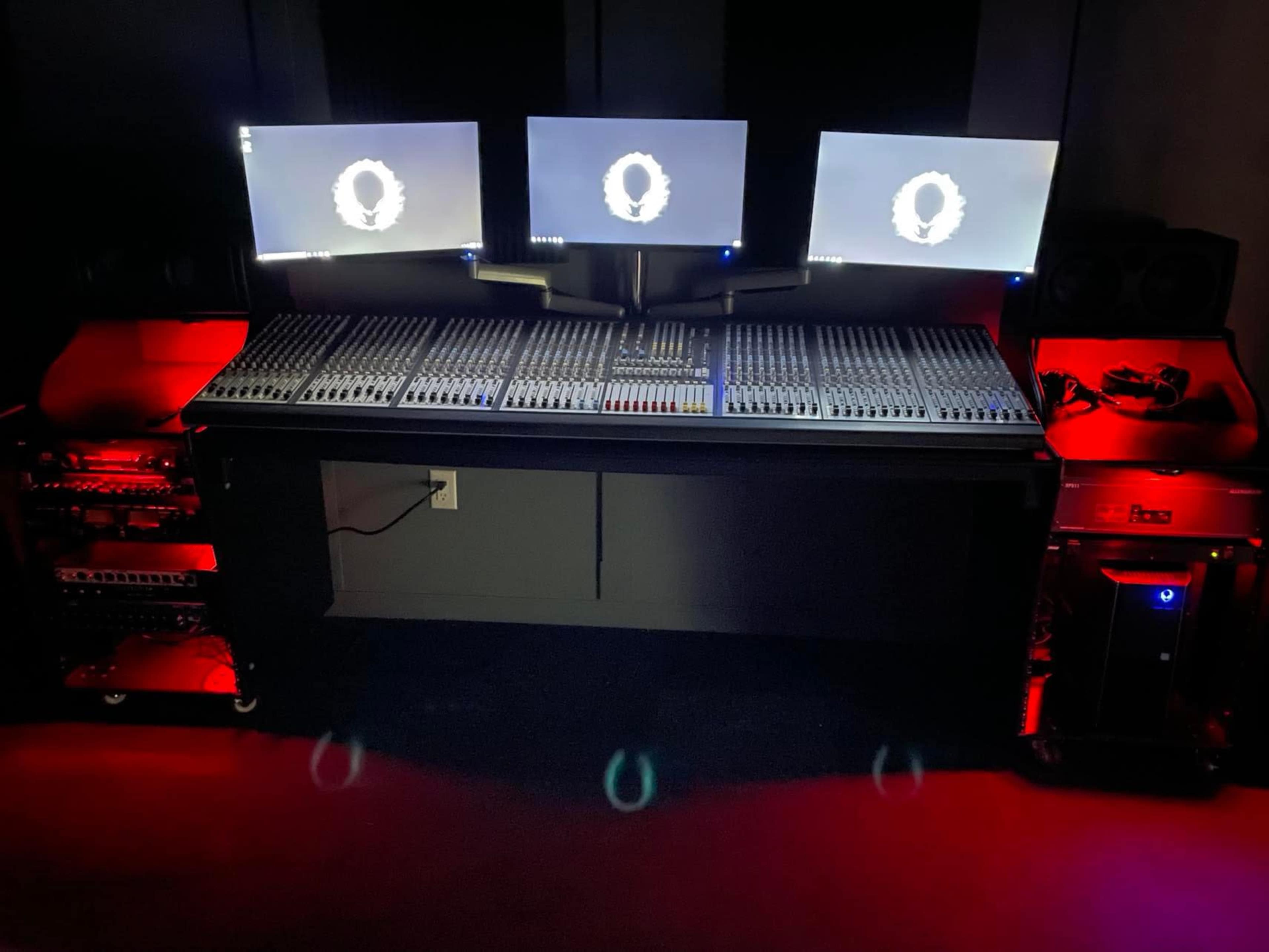 A music studio with a large mixing console, three computer monitors, and illuminated equipment on either side.