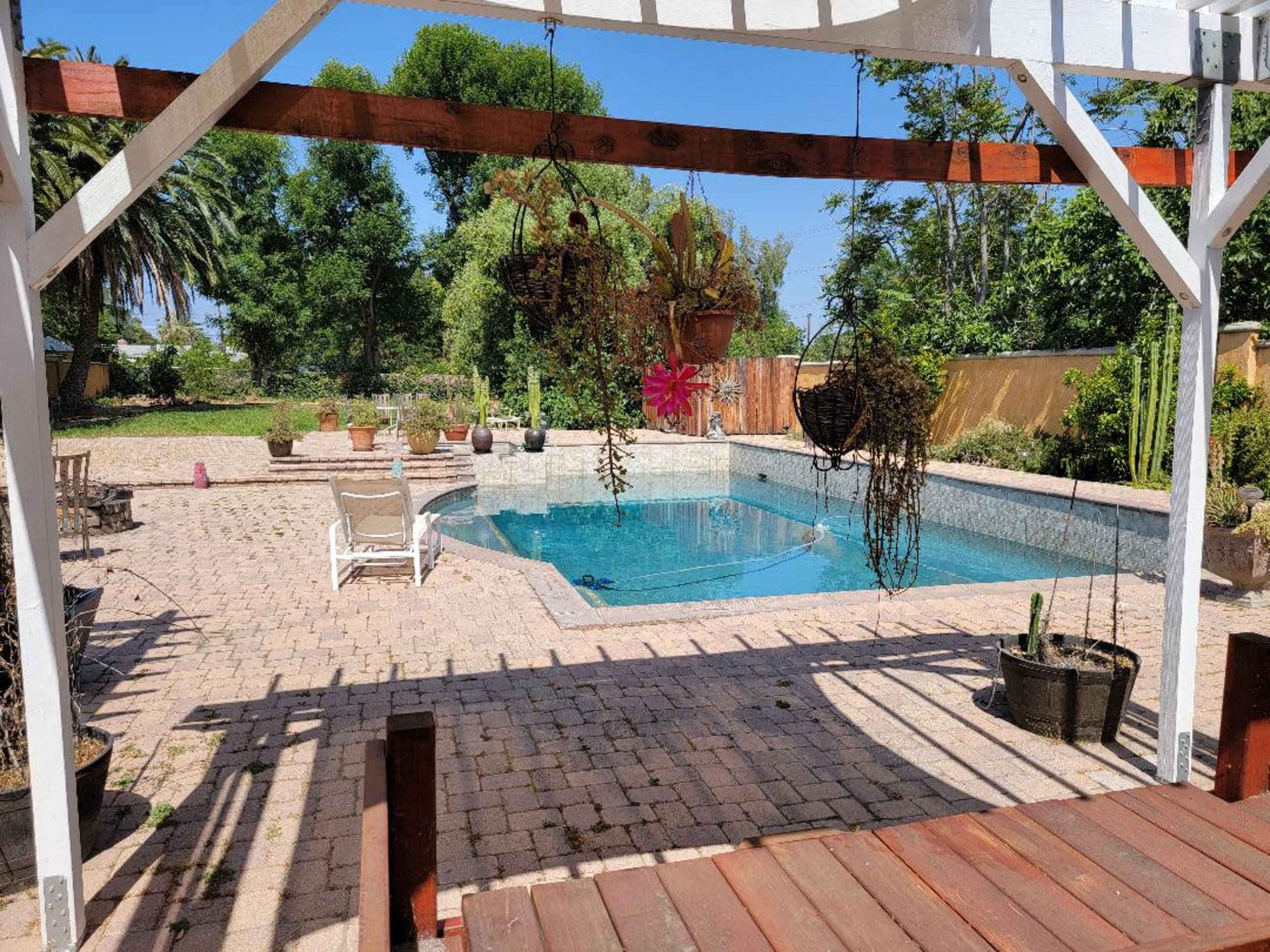 A backyard patio features a swimming pool surrounded by potted plants and sun loungers under a wooden pergola.