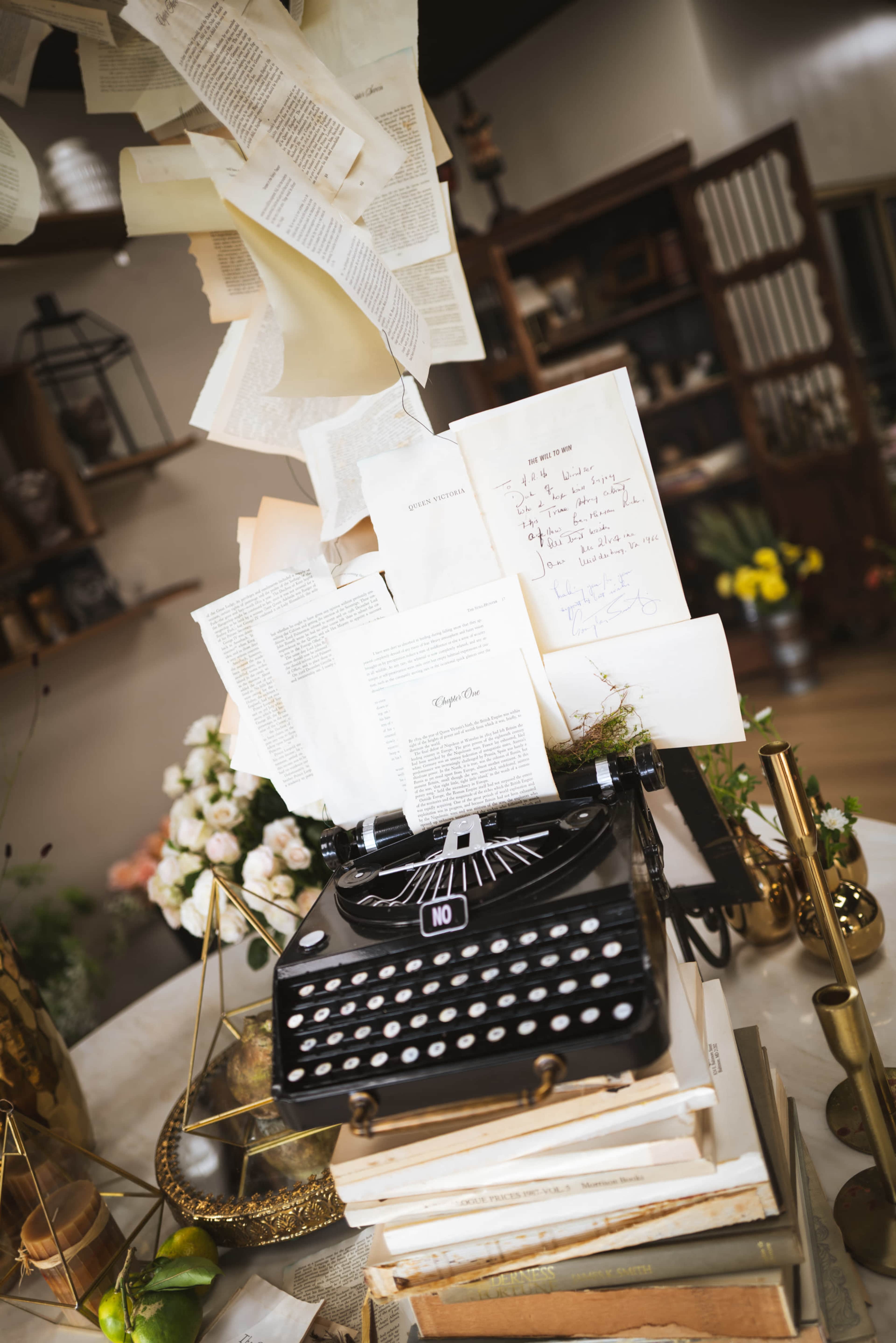 A vintage typewriter is positioned on a stack of books, surrounded by scattered sheets of paper and flowers.