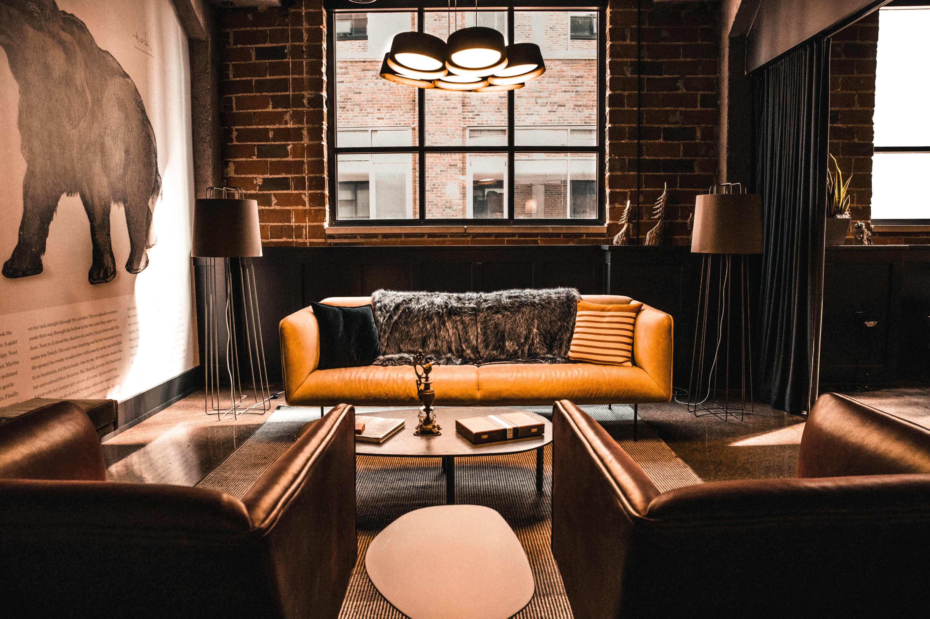 The image shows a modern, cozy living space with a yellow sofa, two brown armchairs, a coffee table, and large windows featuring brick walls.