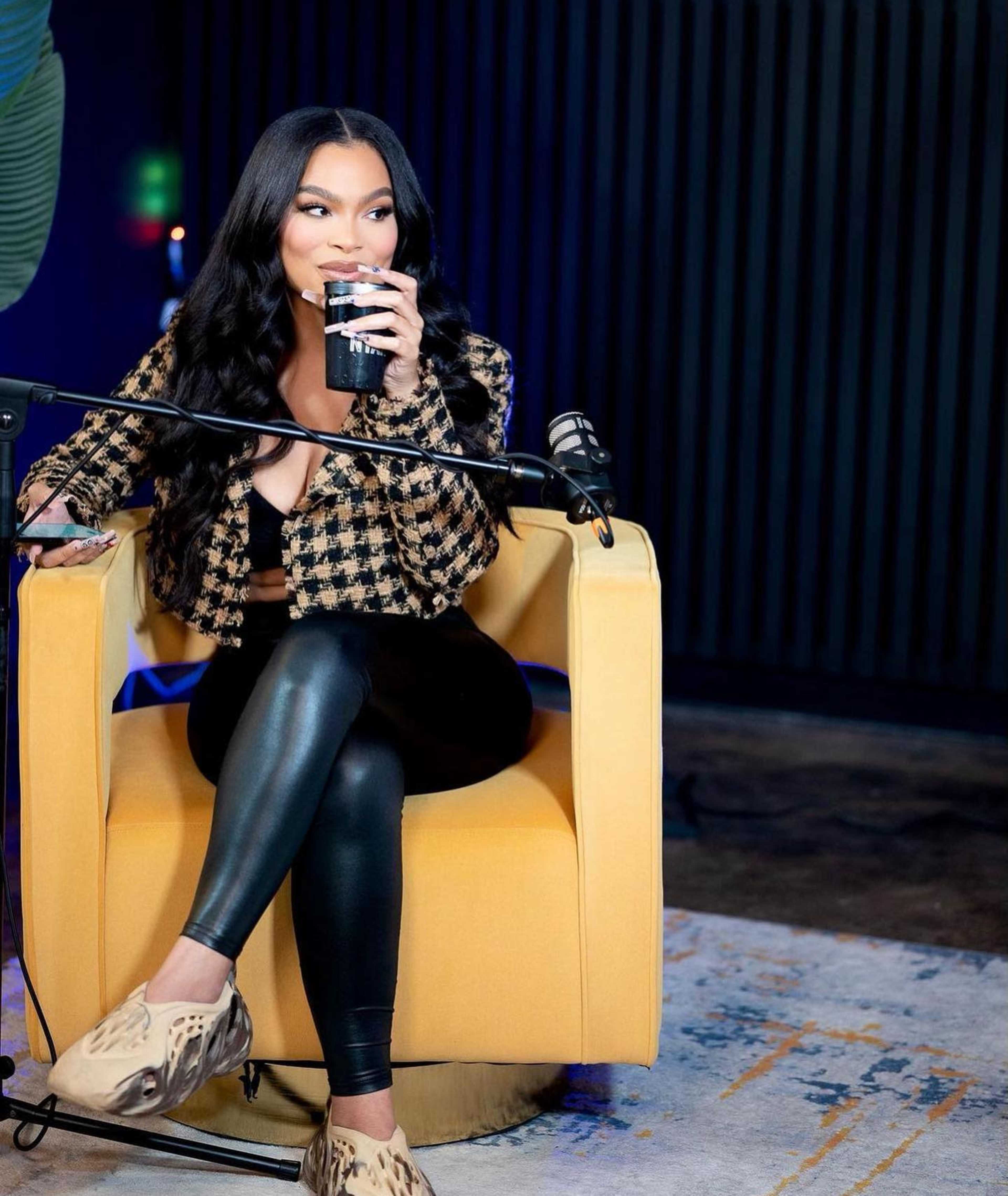 A woman is sitting in a yellow chair, holding a drink while wearing a patterned jacket and black leggings, with a microphone positioned nearby.