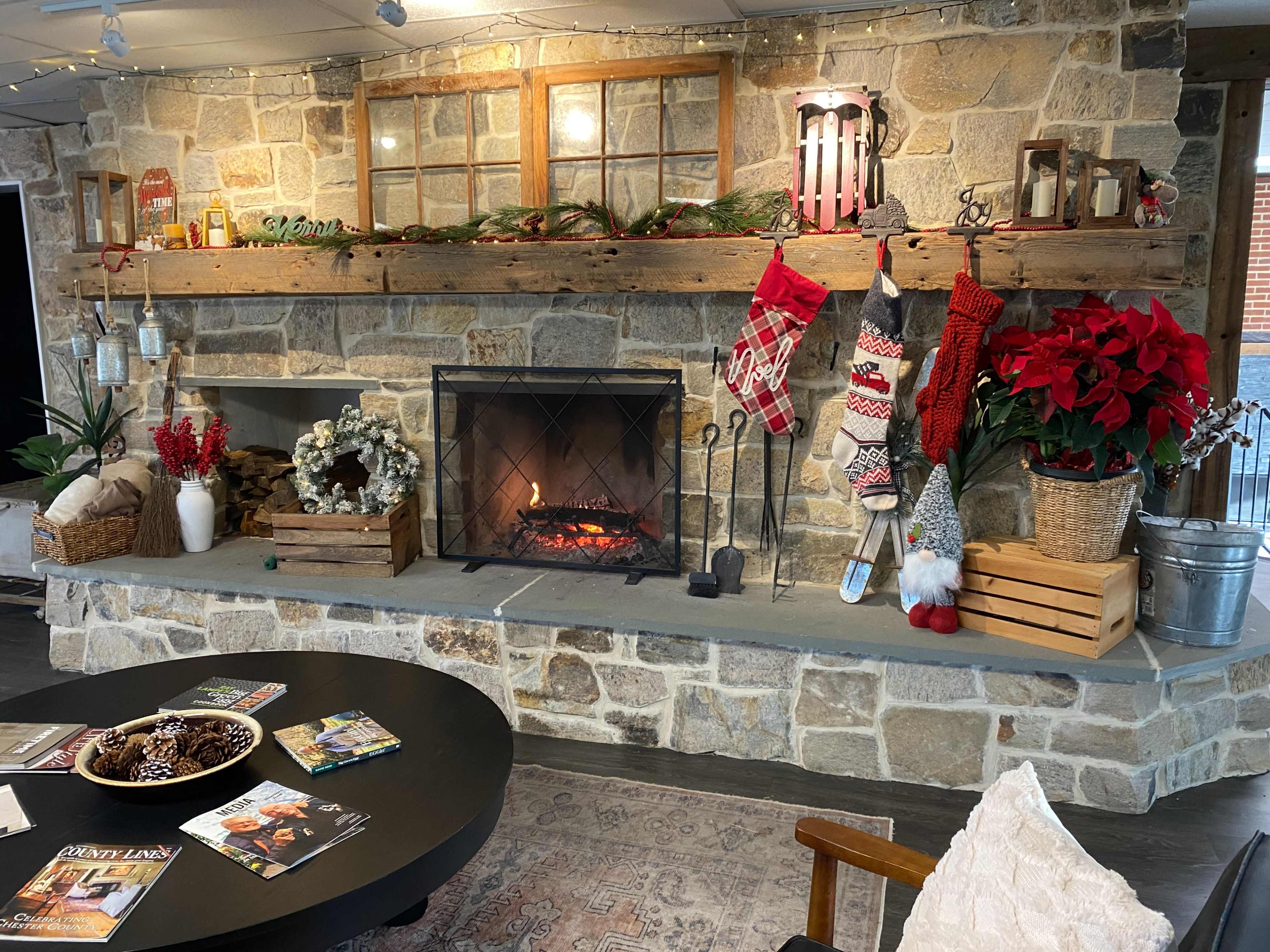 A stone fireplace adorned with holiday decorations, including stockings, garlands, and red poinsettias, while a cozy seating area is visible in front.