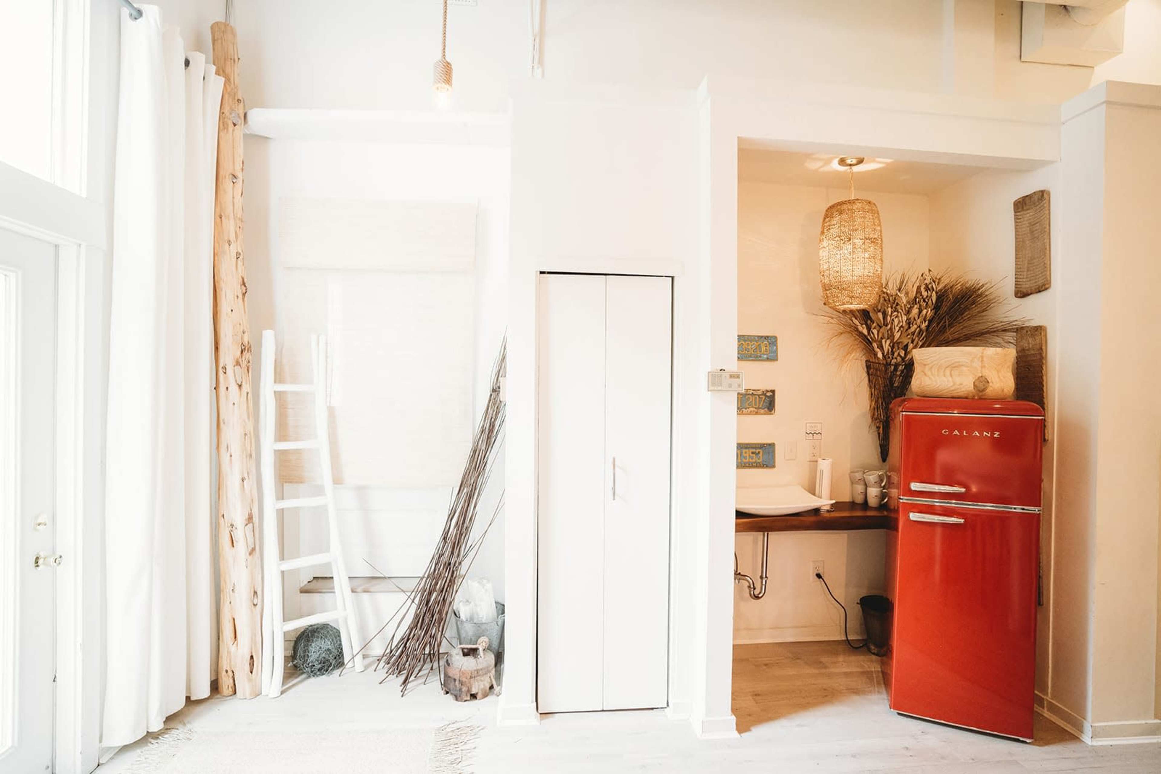 A bright interior space featuring a vintage red refrigerator, a small sink area with woven decor, and light-colored walls with natural elements.