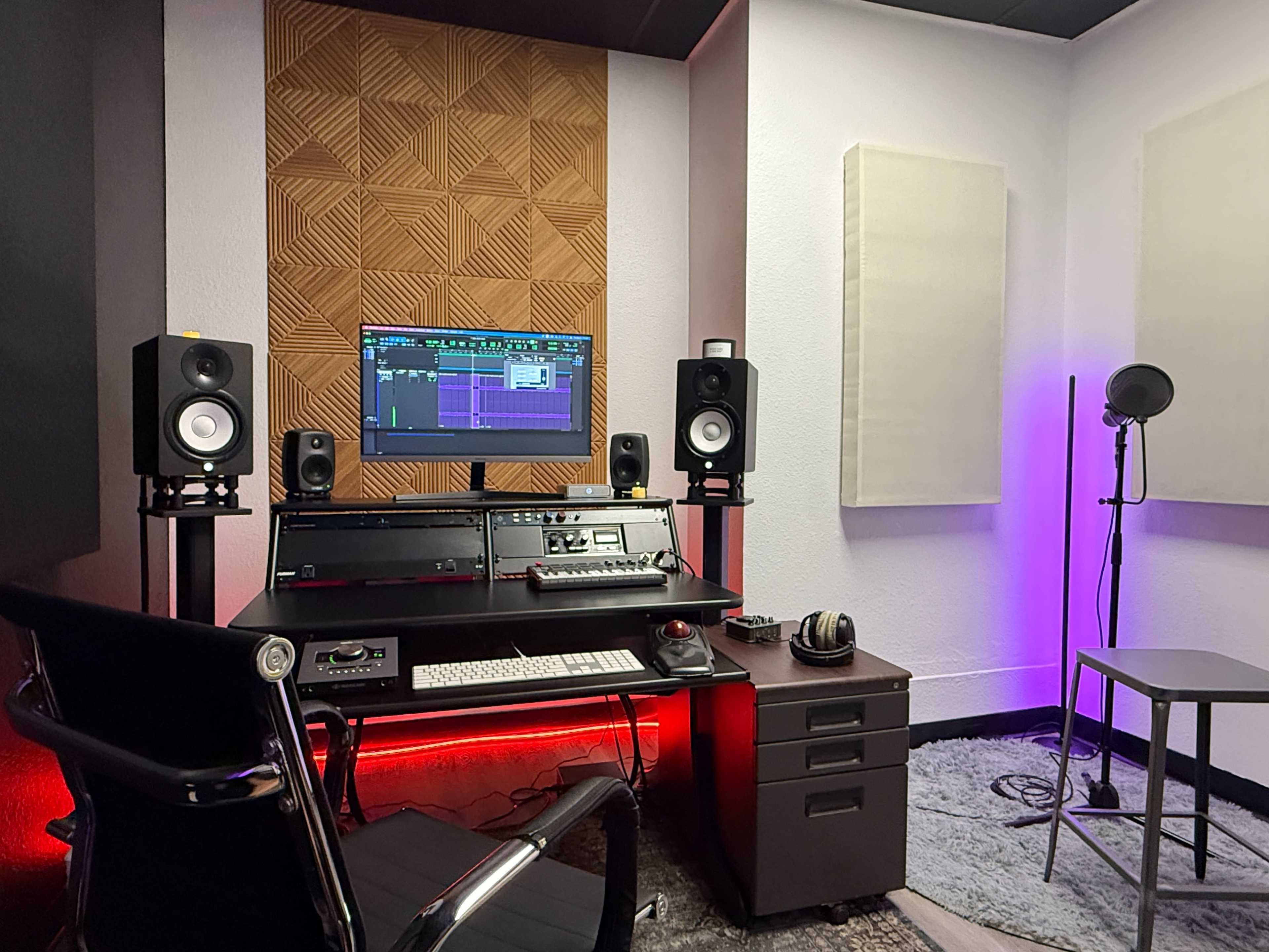 The image shows a music production studio featuring a computer monitor with software, audio speakers, a microphone, and a desk with audio equipment in a sound-treated room.