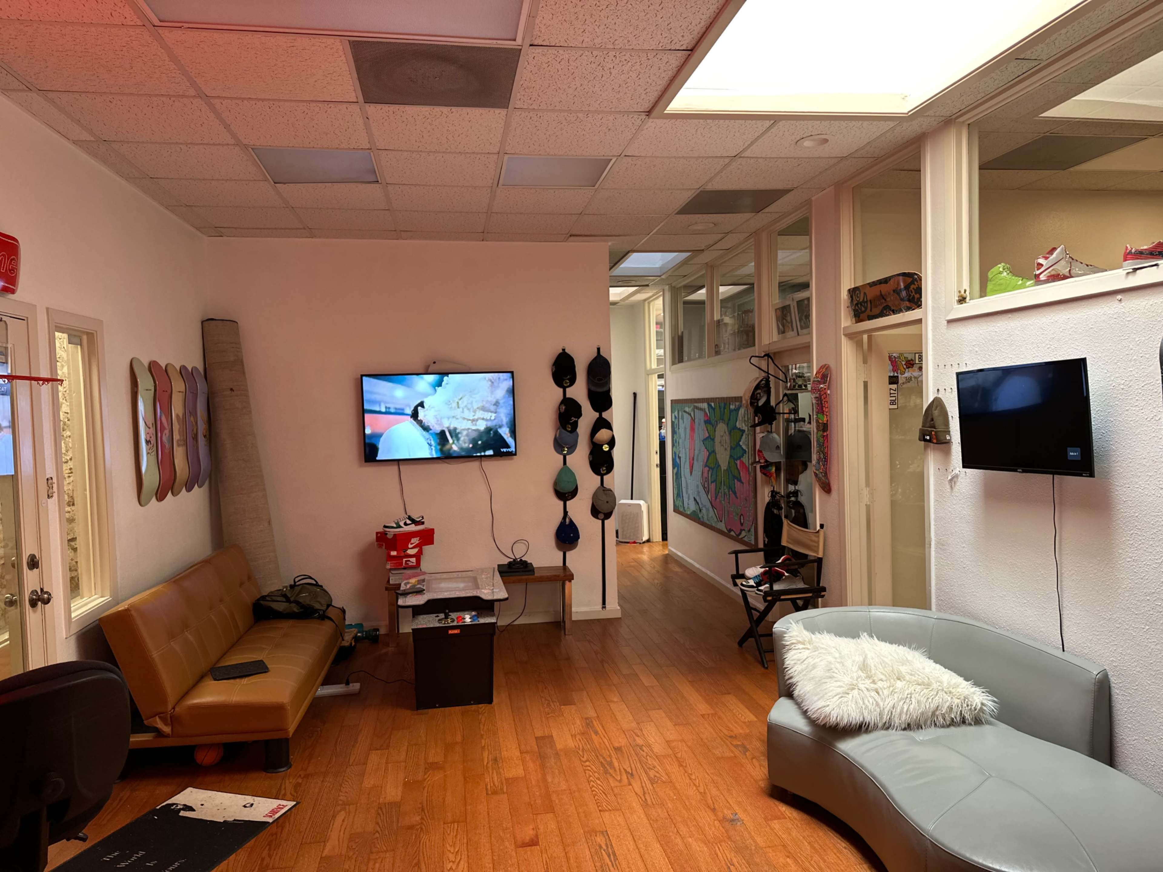 The image shows a spacious indoor area with a brown leather couch, two wall-mounted televisions, and various artworks and skateboards displayed along the walls.