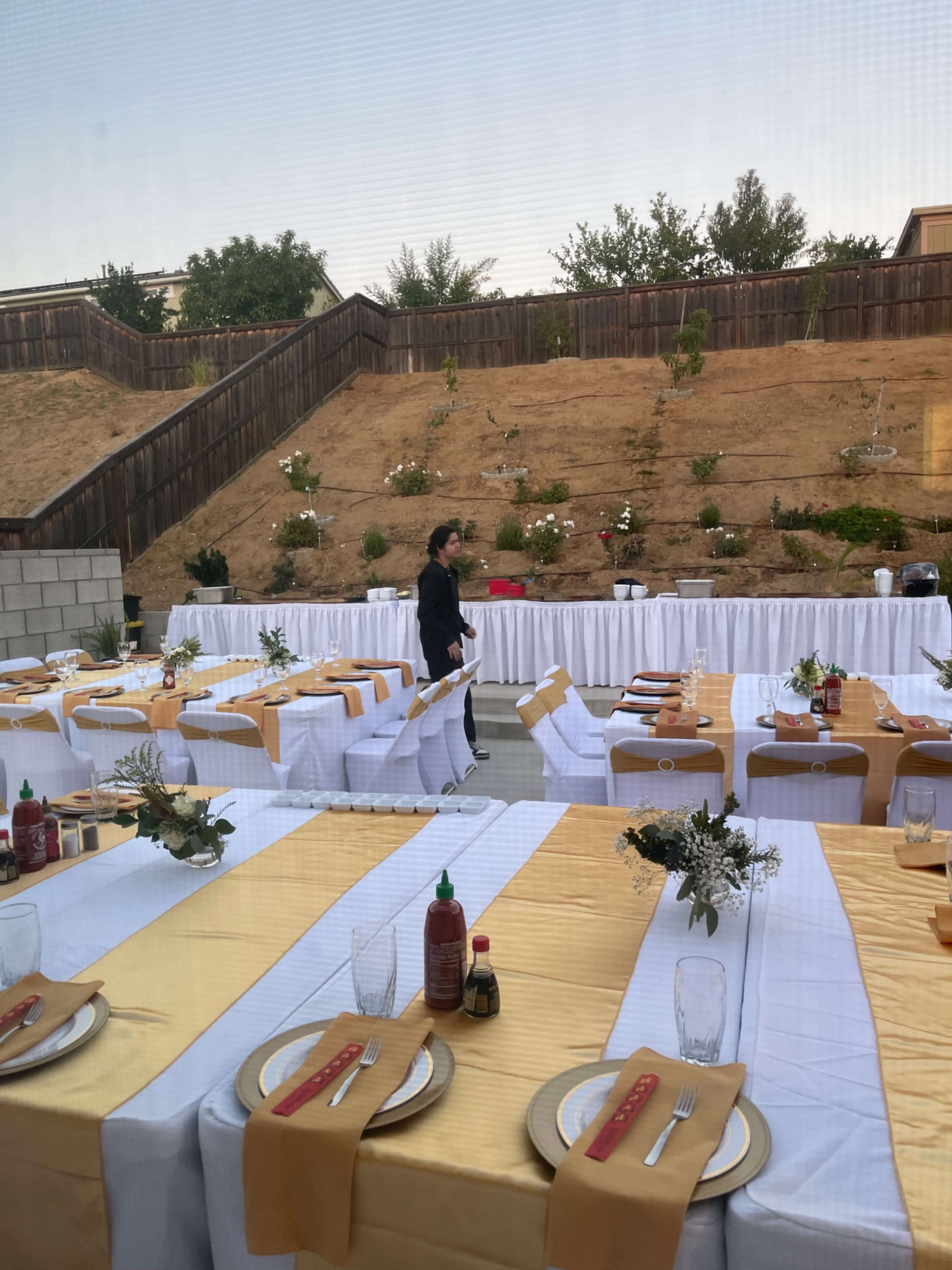 A person is walking past a long table setup with white and gold table settings in a yard featuring landscaping in the background.
