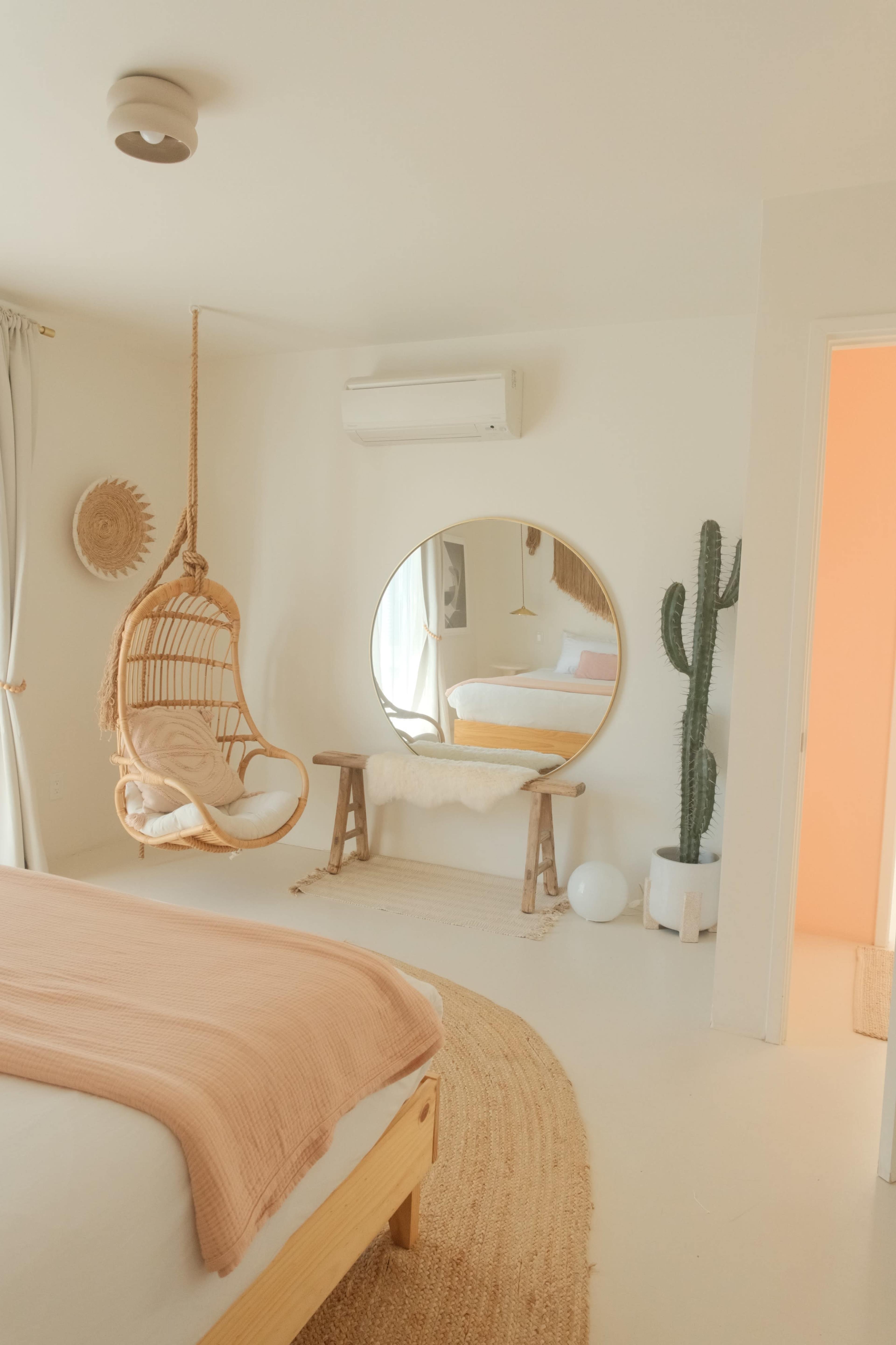 The image shows a bright, minimalist bedroom featuring a hanging chair, a round mirror, a cactus, and a soft rug on the floor.