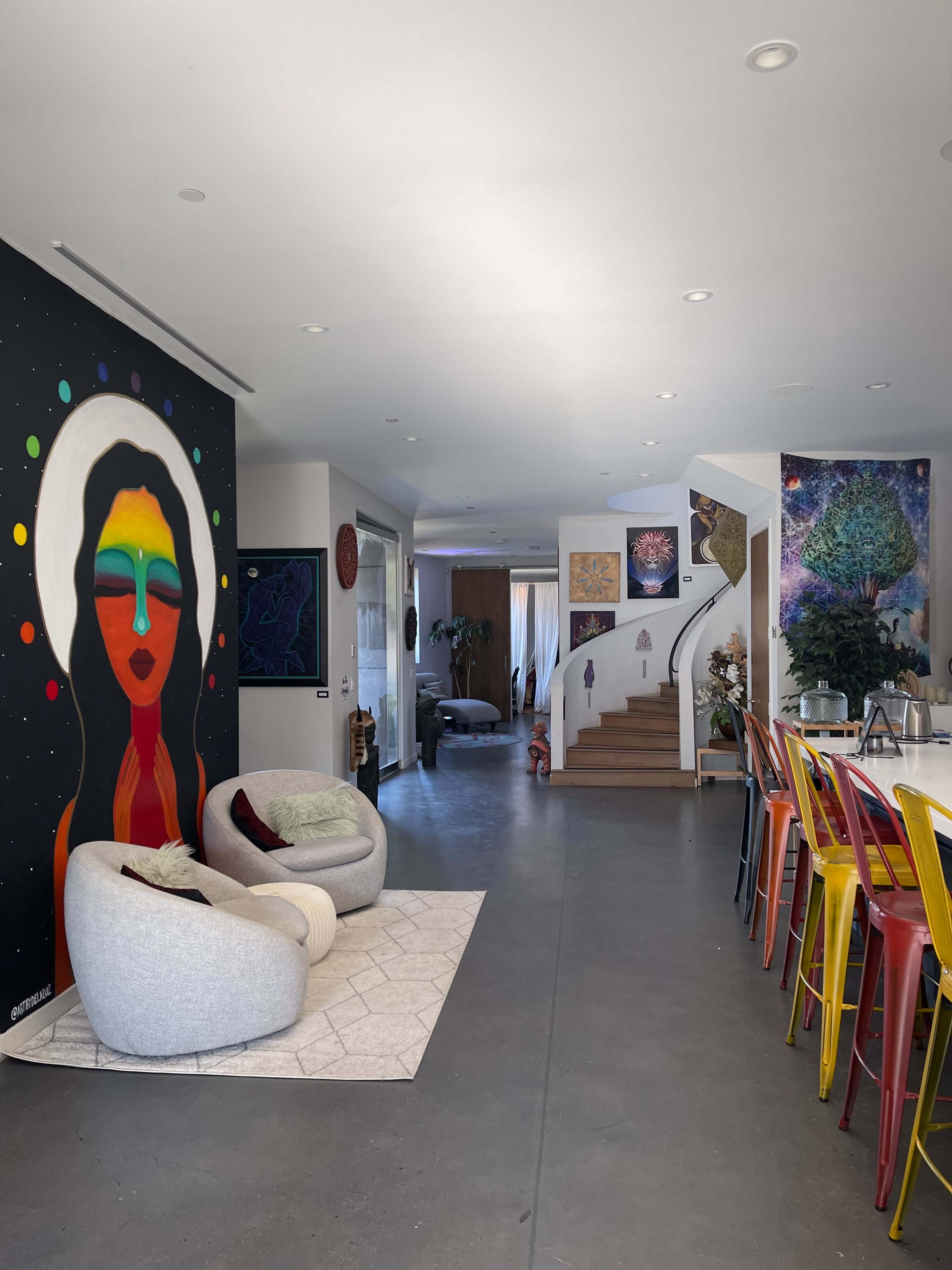 A modern, open-concept living space with a colorful mural of a woman on the wall, cozy seating near the entrance, and a dining area with vibrant, multicolored chairs.