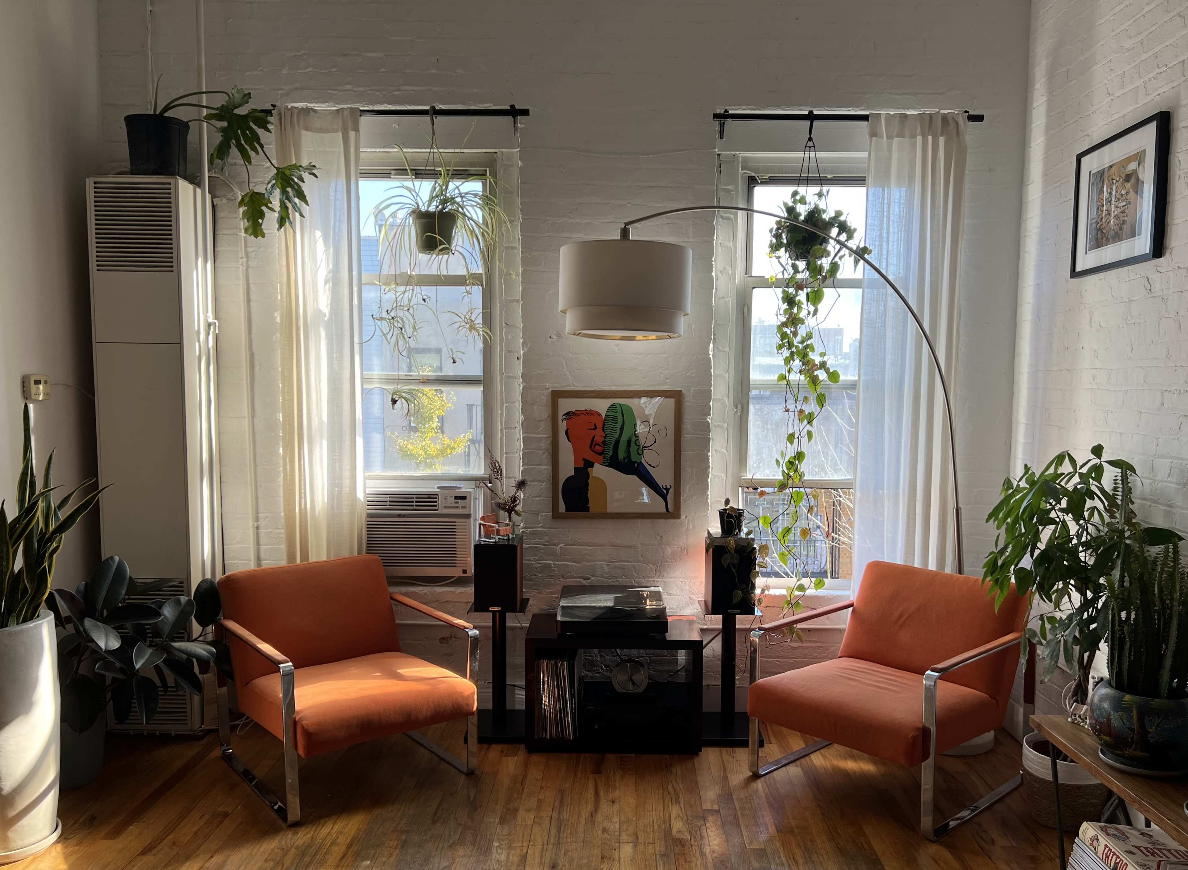 The image shows a bright living room with two orange chairs facing a coffee table, flanked by large windows with plants and a wall-mounted lamp.