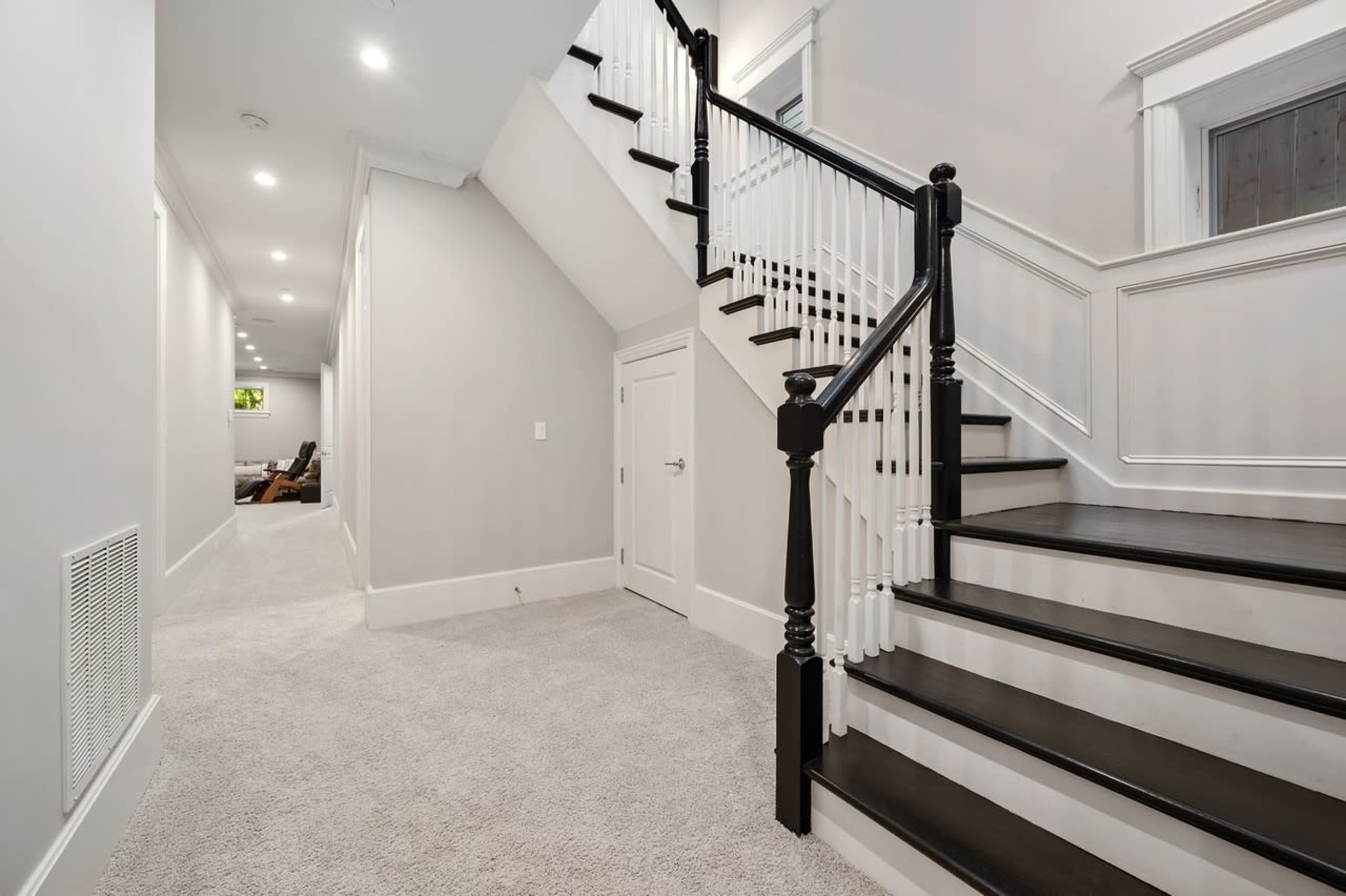 A well-lit hallway leading to a staircase with black railing and carpeted flooring.