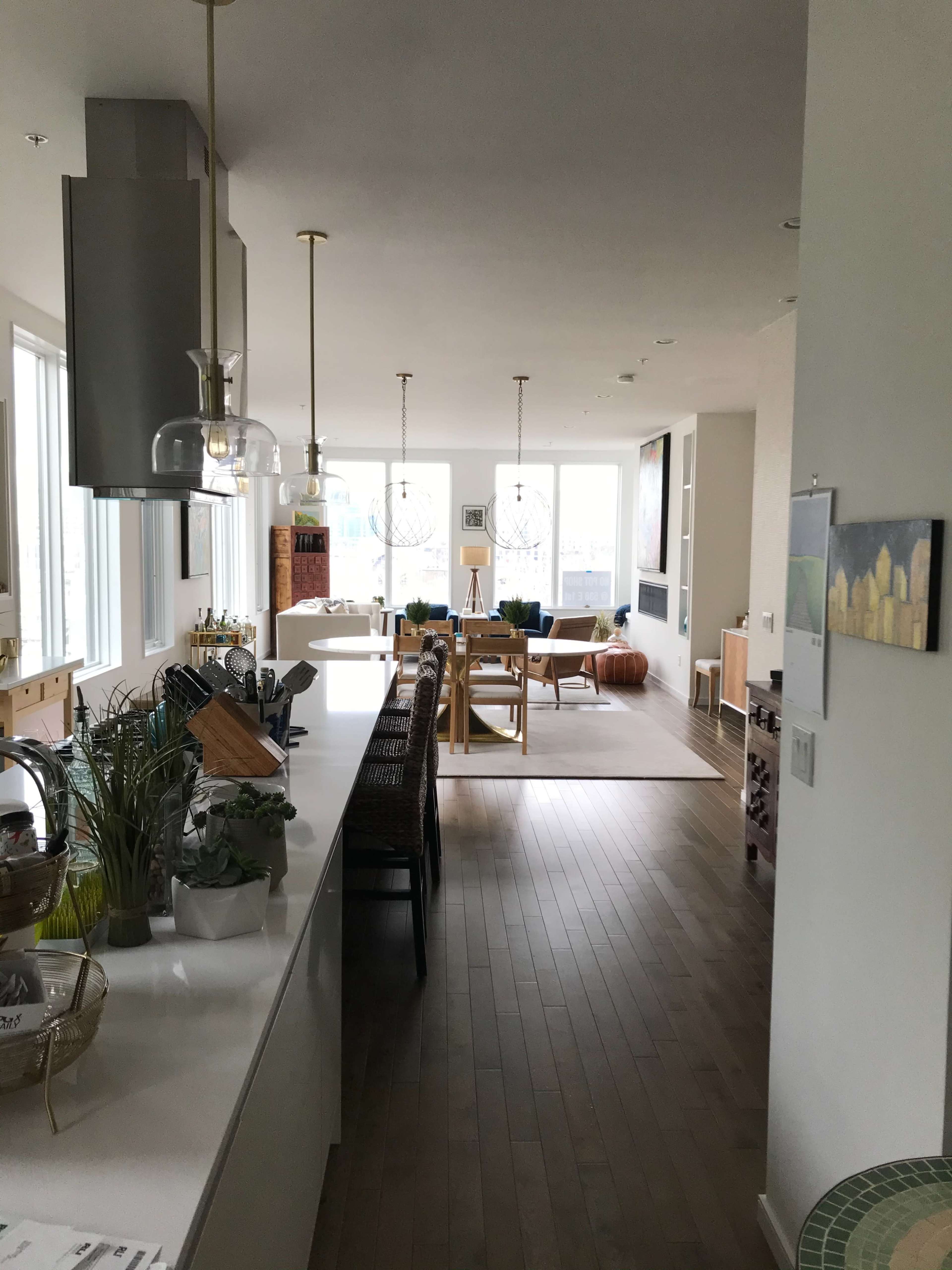 The image shows a modern kitchen and living area with large windows, wooden flooring, and minimalistic furniture.