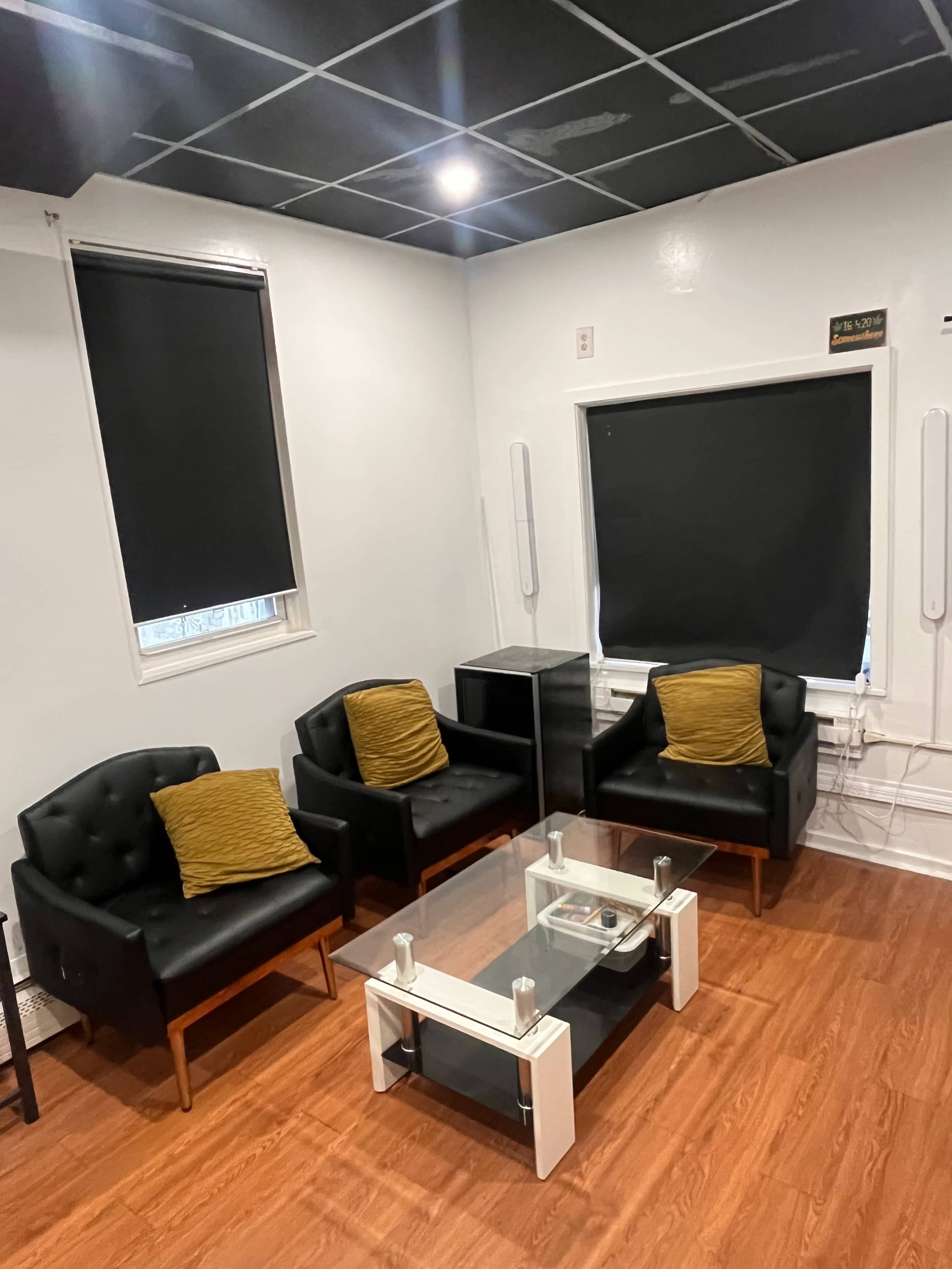 The image shows a small lounge area with three black armchairs, two yellow cushions, a glass coffee table, and a black cabinet, all situated on a wooden floor.