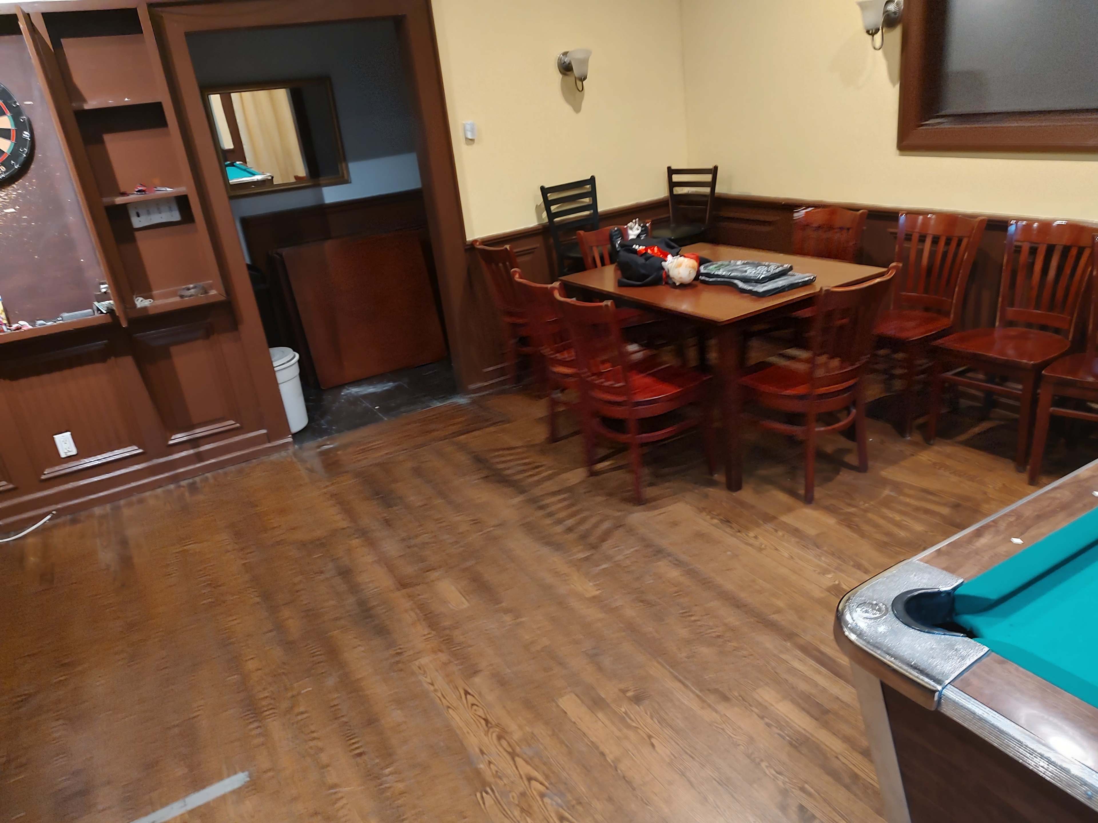 The image shows a room with wooden flooring, a pool table in the foreground, and a dining area with a table and several wooden chairs in the background.