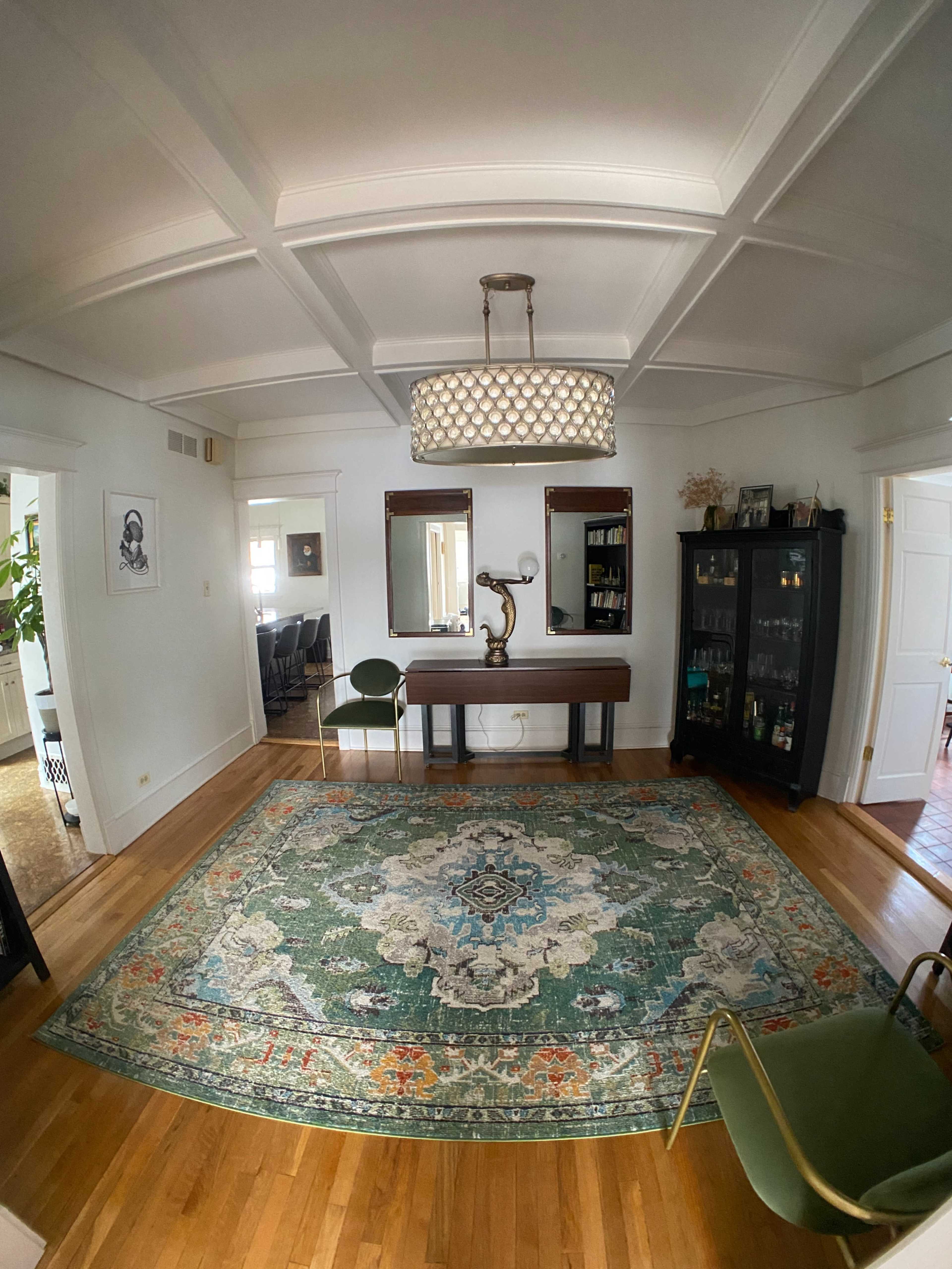 A spacious room with a patterned area rug, a wooden console table with a lamp, two chairs, and a large mirror on the wall.