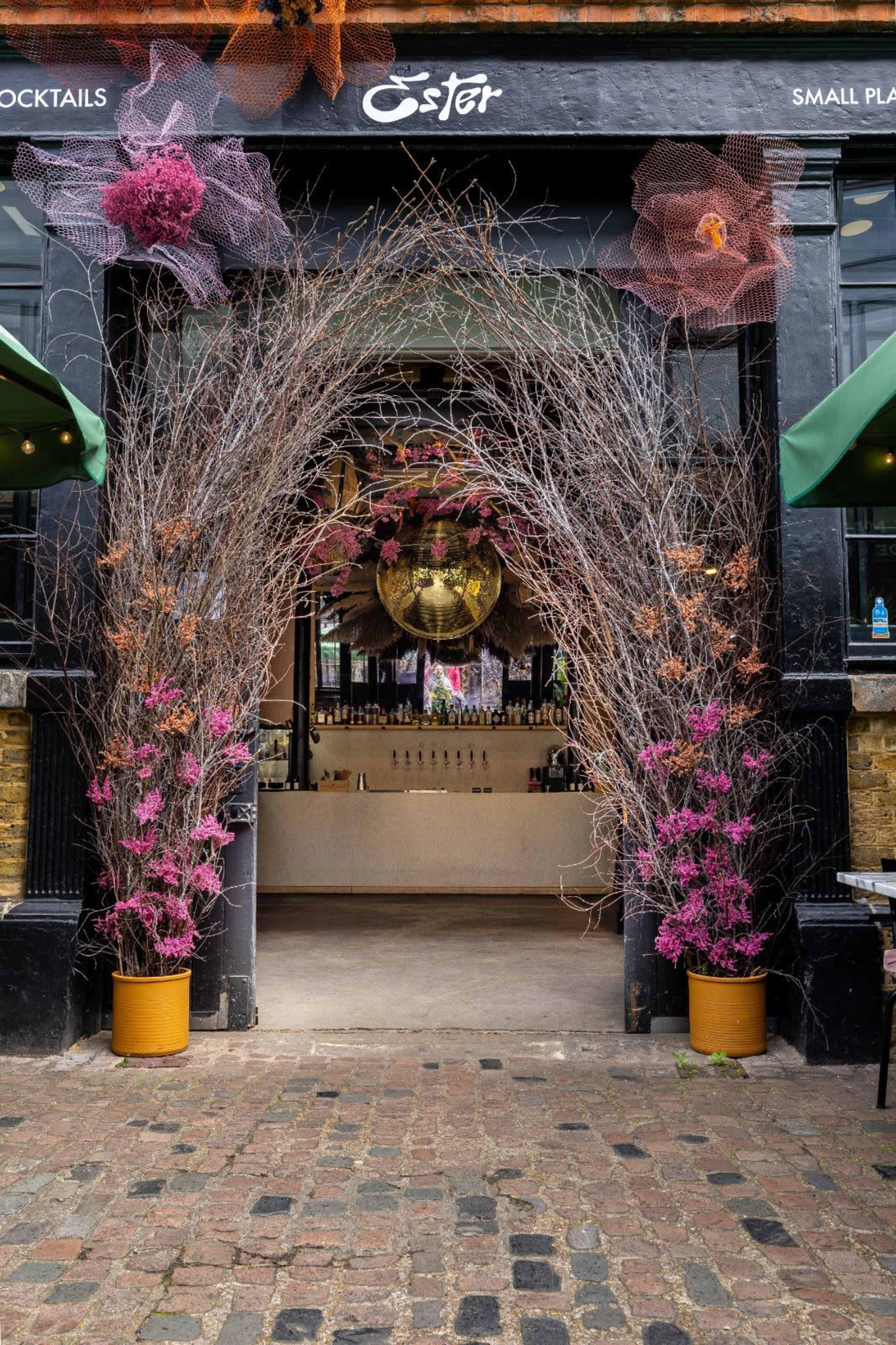 The entrance to a bar features an arch of bare branches adorned with colorful flowers, leading to an interior space with a large disco ball and a bar area.