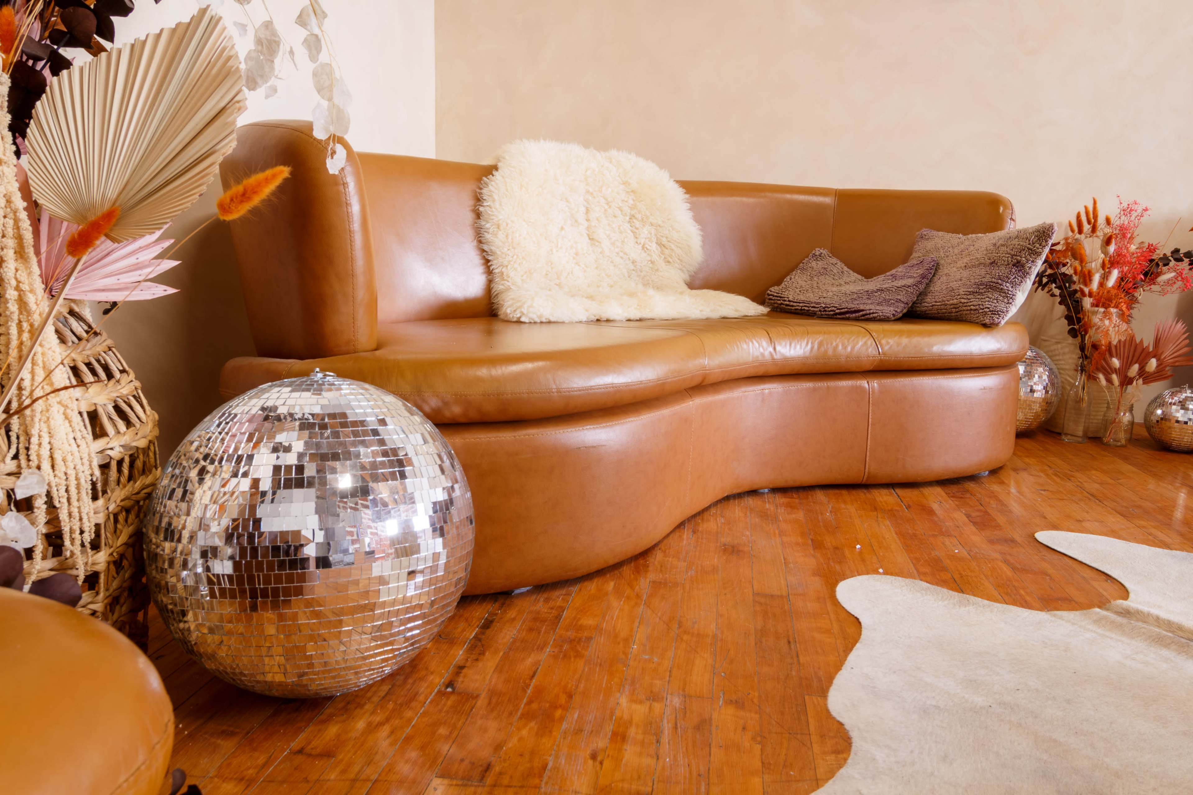 A curved brown leather couch adorned with pillows and a fluffy throw, next to a large disco ball and decorative plants in a warmly lit room.