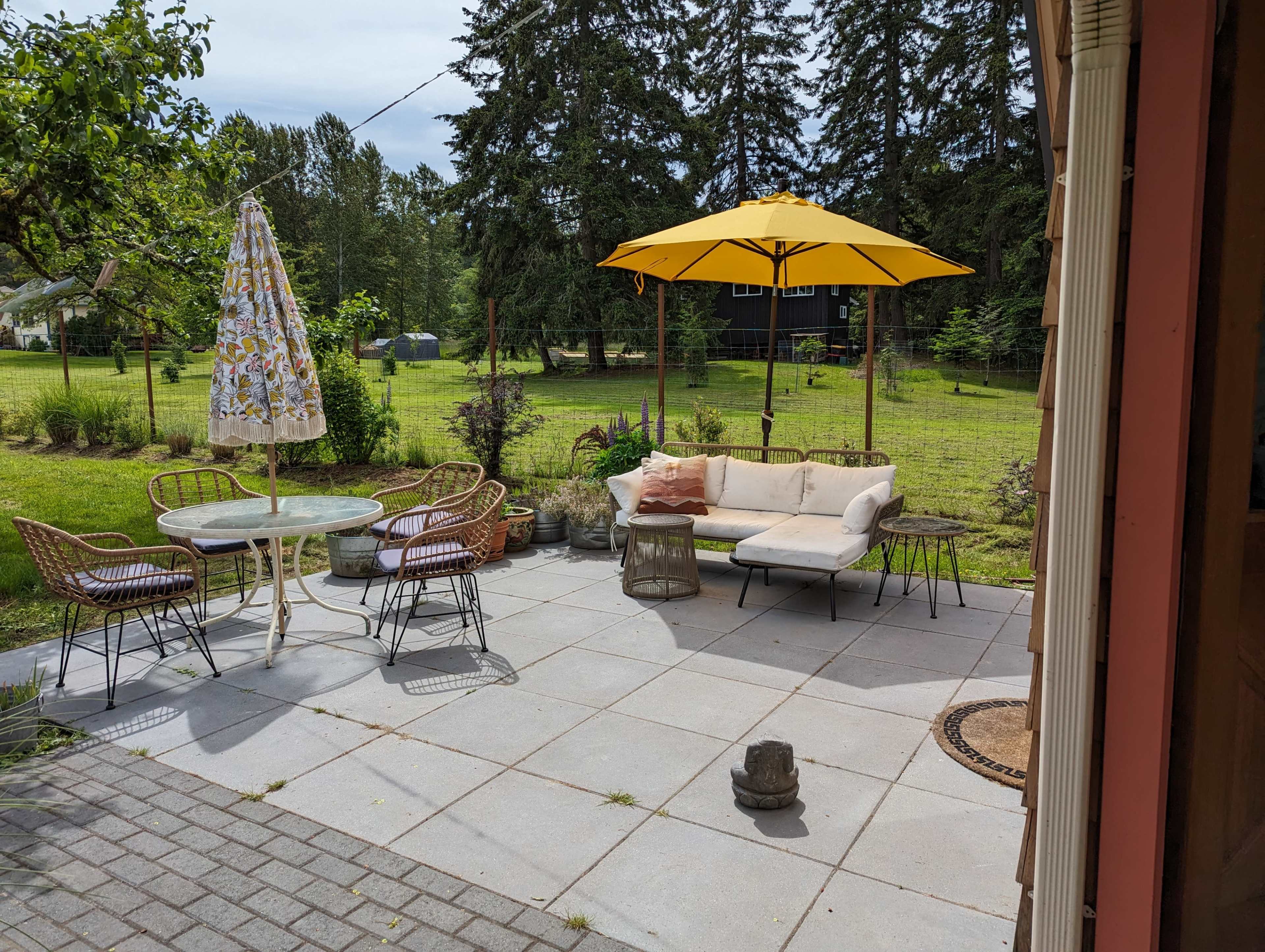 A patio area features a yellow umbrella over a seating arrangement, a table, and chairs surrounded by greenery.