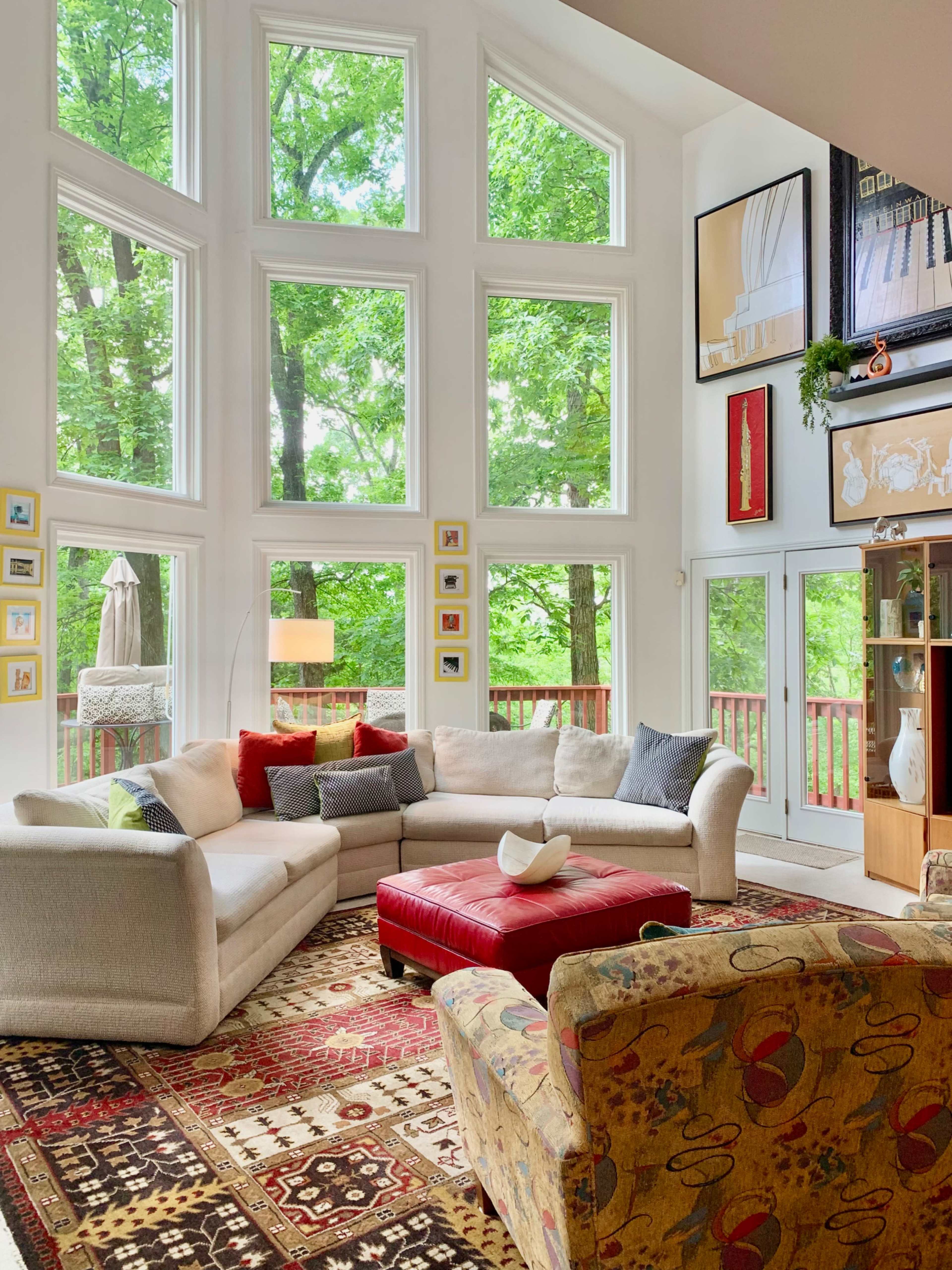 A spacious living room features large windows that overlook a green landscape, with a sectional couch, a red ottoman, and a patterned armchair arranged around a decorative rug.