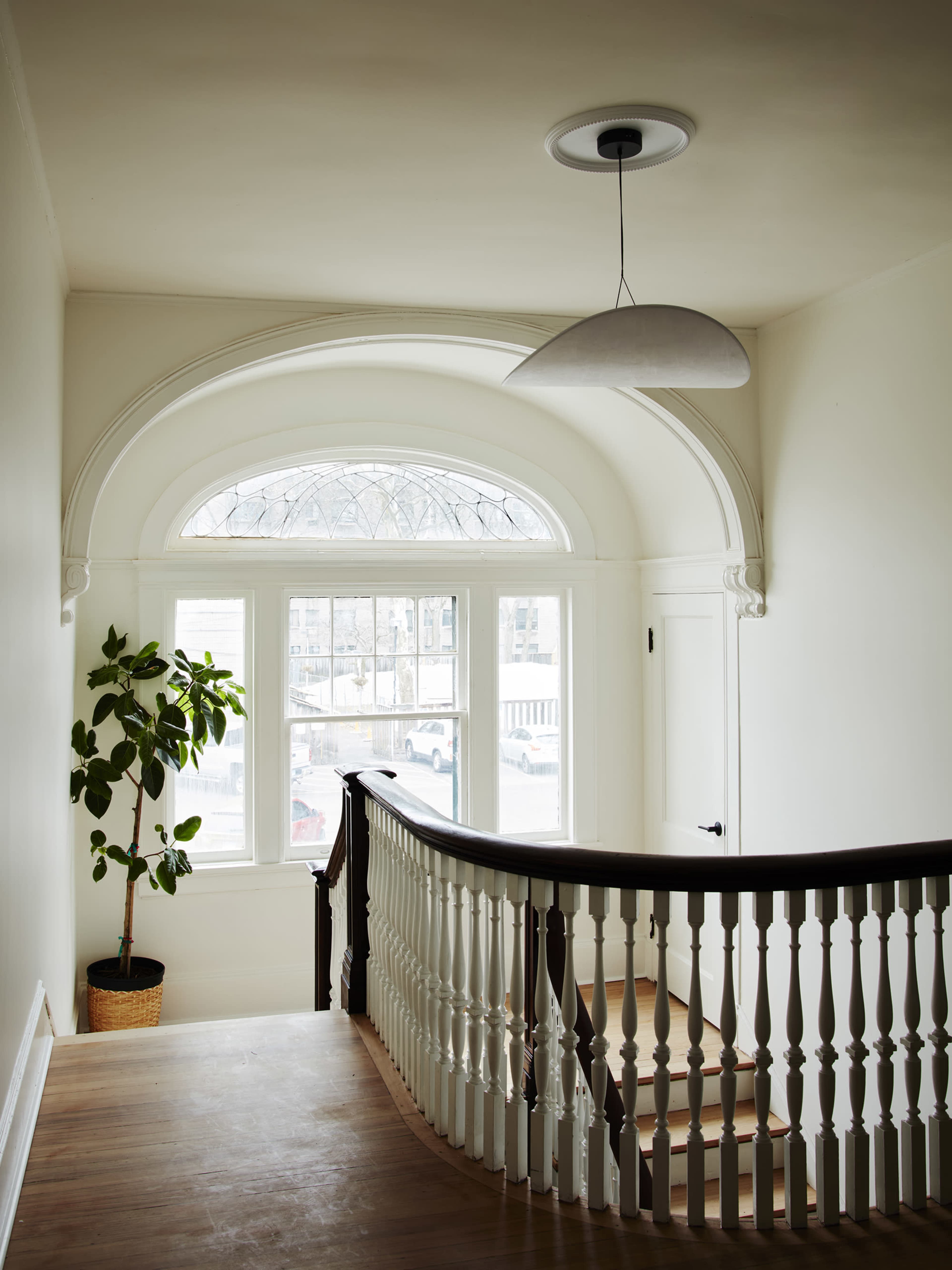 A staircase with a wooden banister leads to a landing featuring a large arched window and a potted plant.