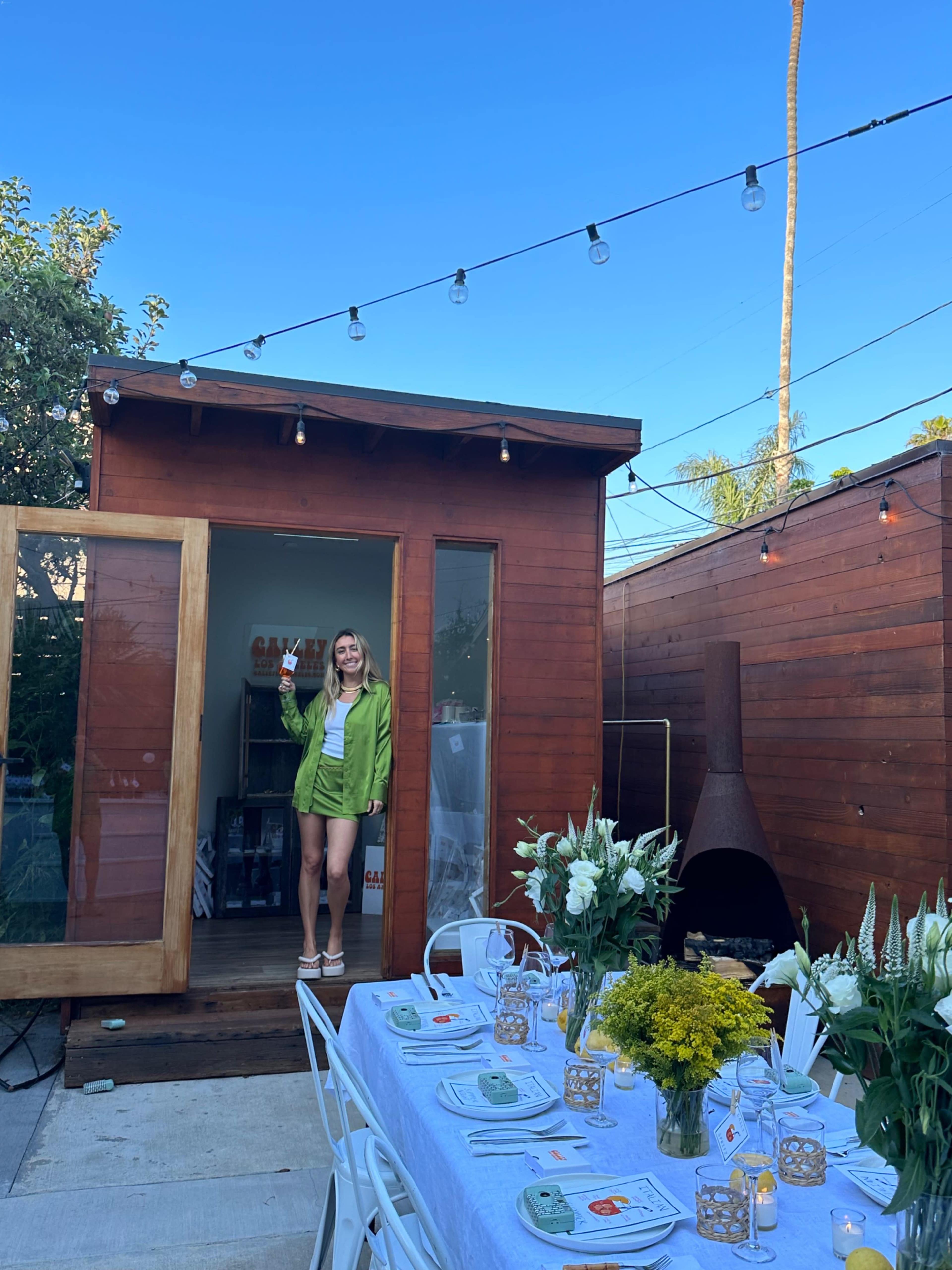 A woman in a green jacket stands at the entrance of a wooden structure, with a dining table set nearby under string lights.