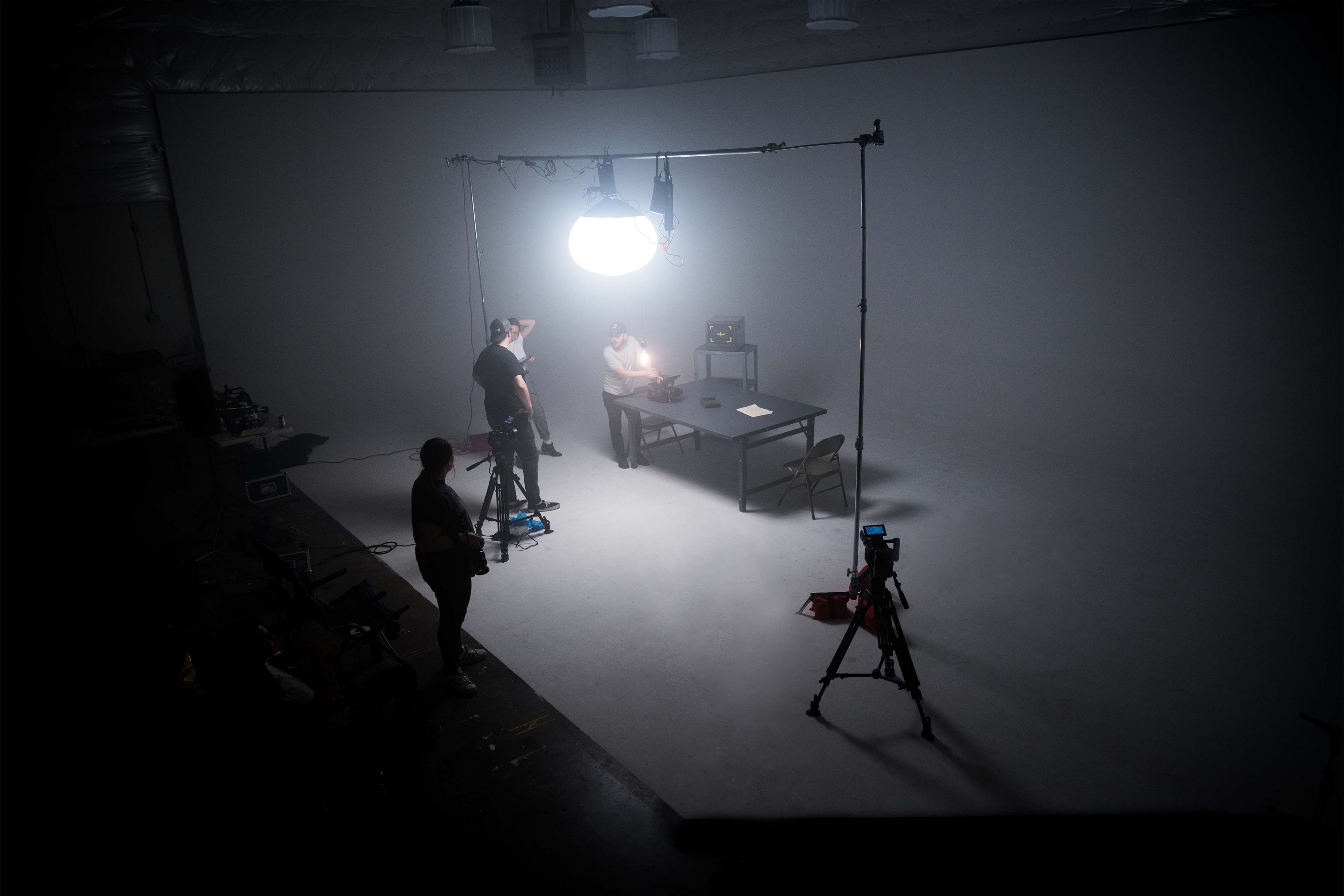 A brightly lit studio setup shows a table with a person seated, surrounded by cameras and crew members in the background.