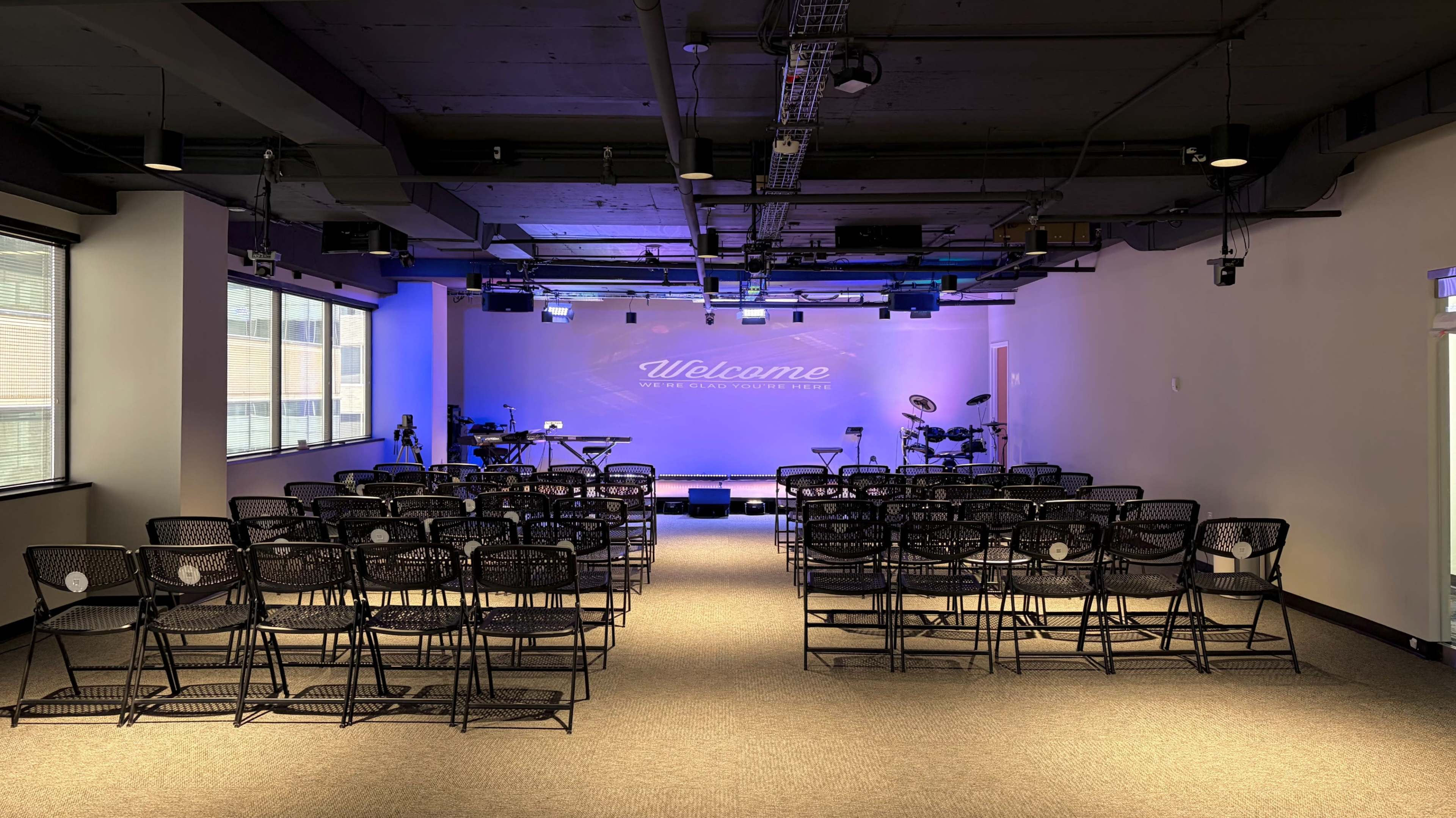 The image shows an empty event space with rows of black chairs arranged facing a stage that has band instruments and purple lighting.