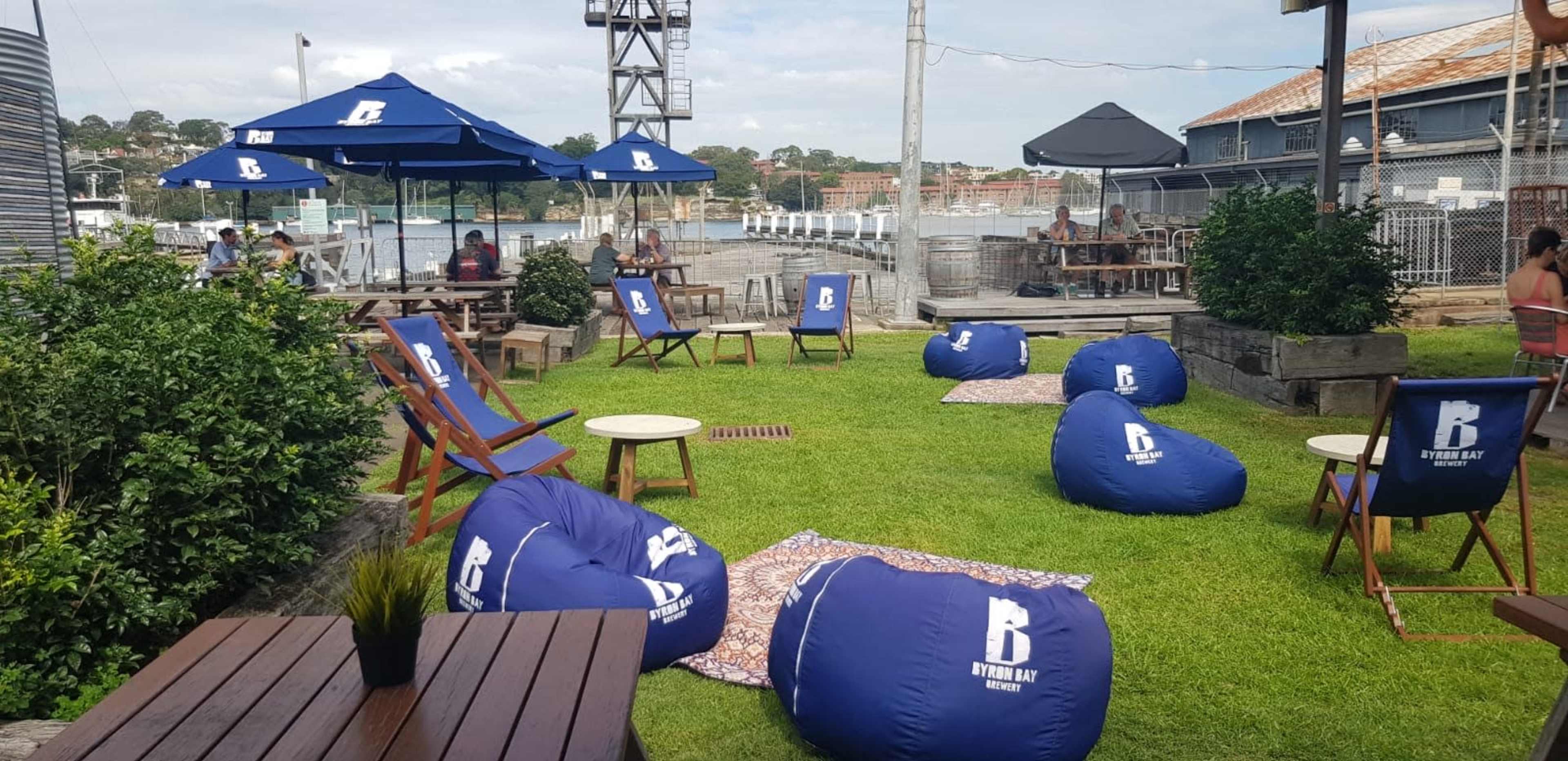 A outdoor seating area with bean bags, wooden tables, and umbrellas, set in a grassy space near a waterfront.